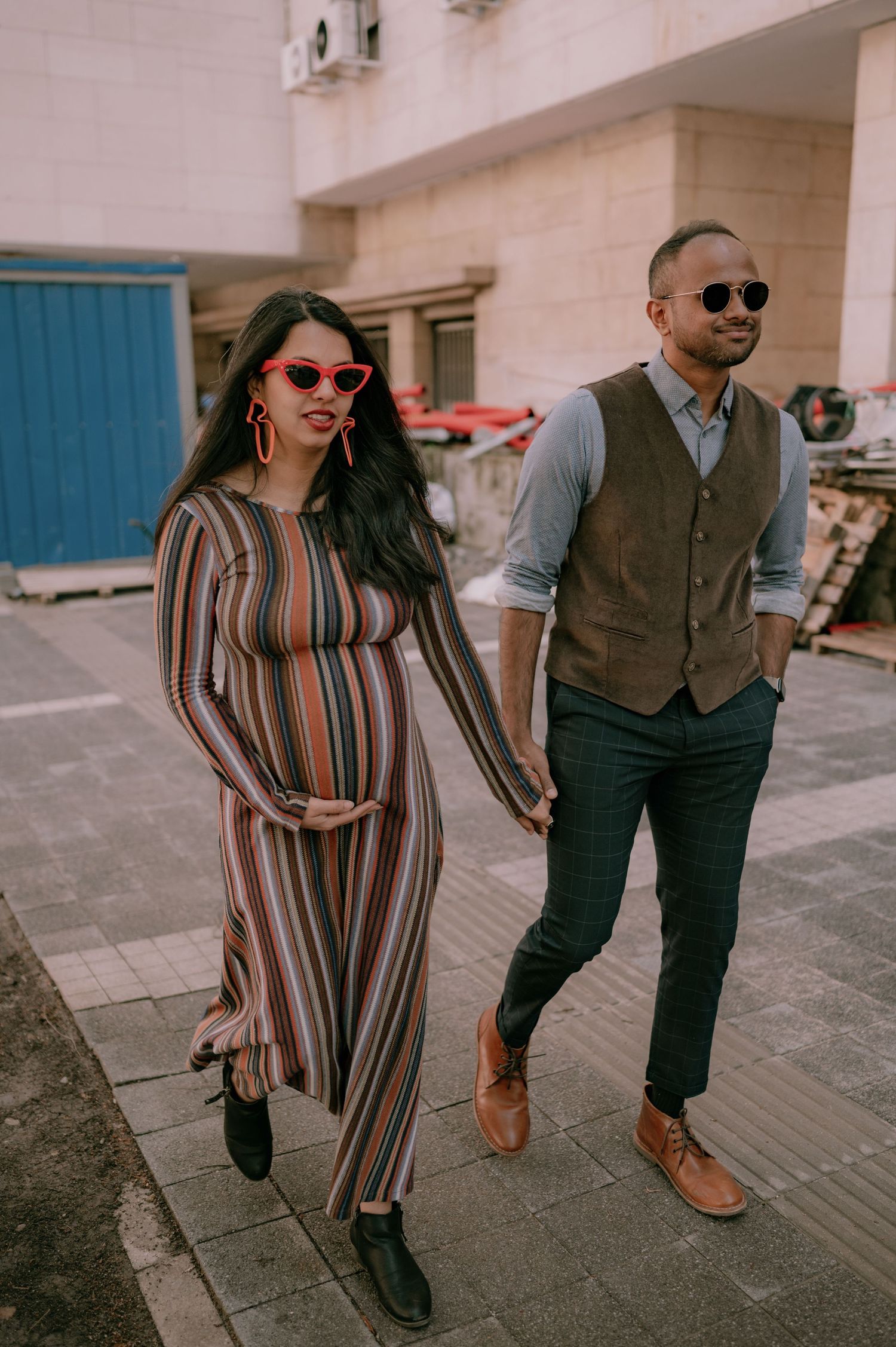Pregnant woman in striped dress and red sunglasses walks hand-in-hand with stylish man in vest outdoors.