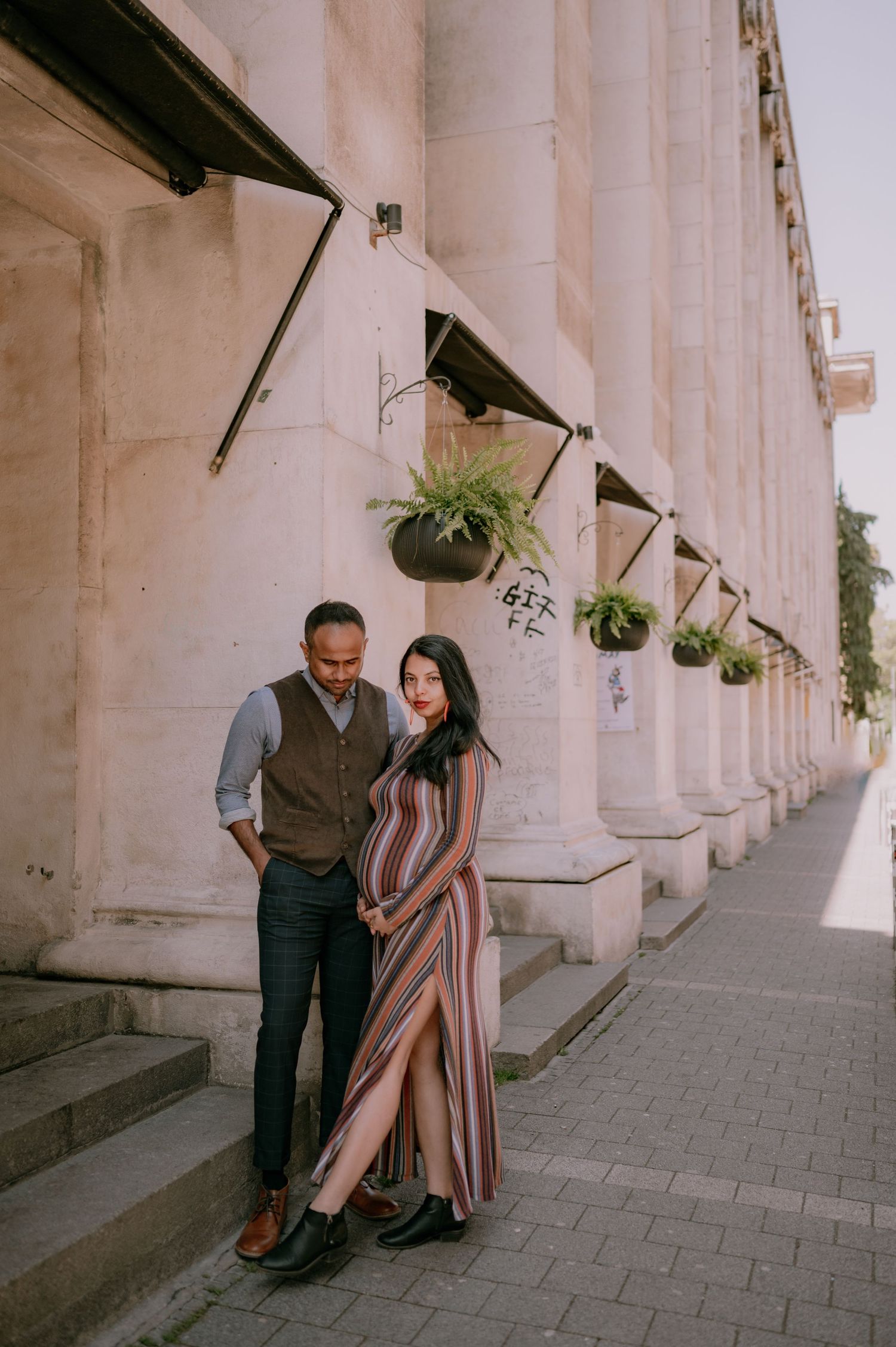Pregnant couple posing beside elegant stone columns with hanging ferns on a sunny urban sidewalk.