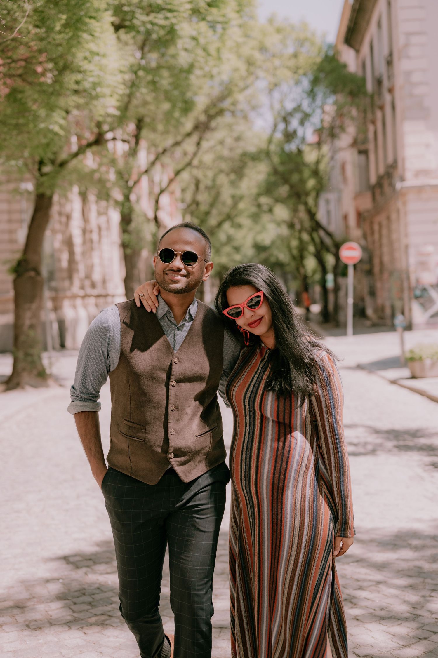 A stylish couple wearing sunglasses poses on a cobblestone street, man in vest and woman in striped dress.