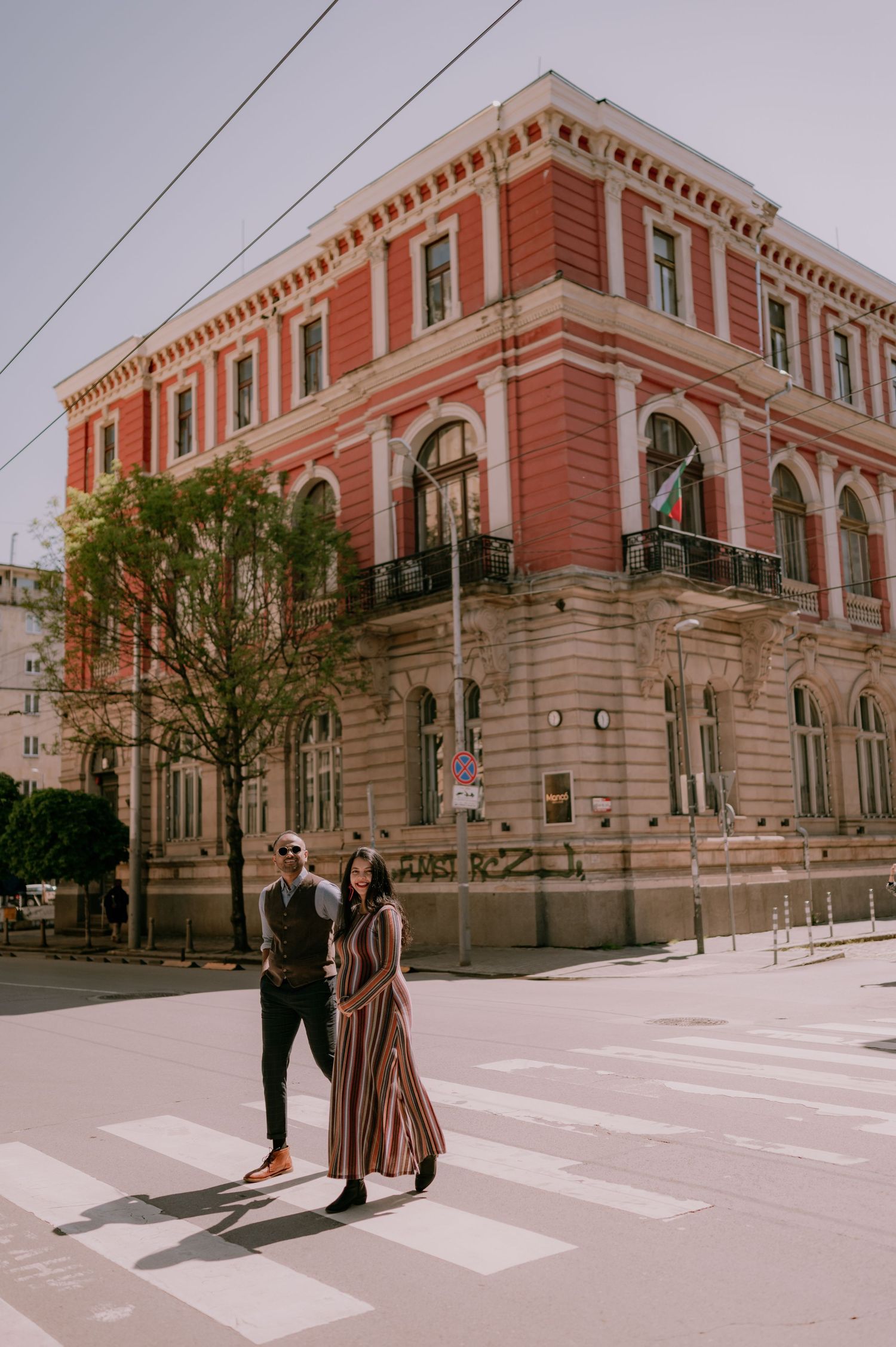 Couple crossing a zebra crossing in front of a grand red historic building on a sunny urban street.
