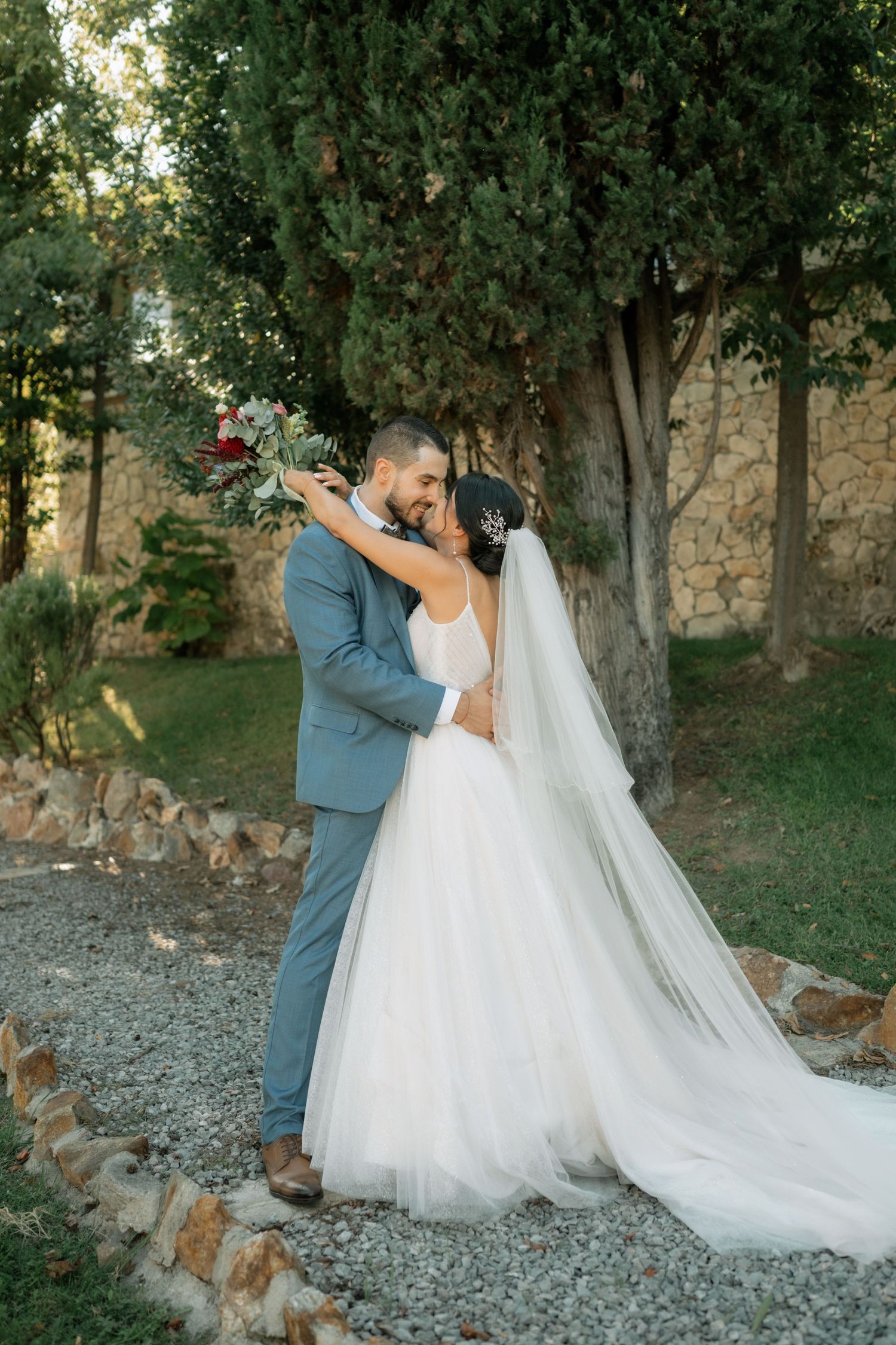 A couple in wedding attire share an intimate moment on a rocky outdoor path surrounded by trees at sunset.