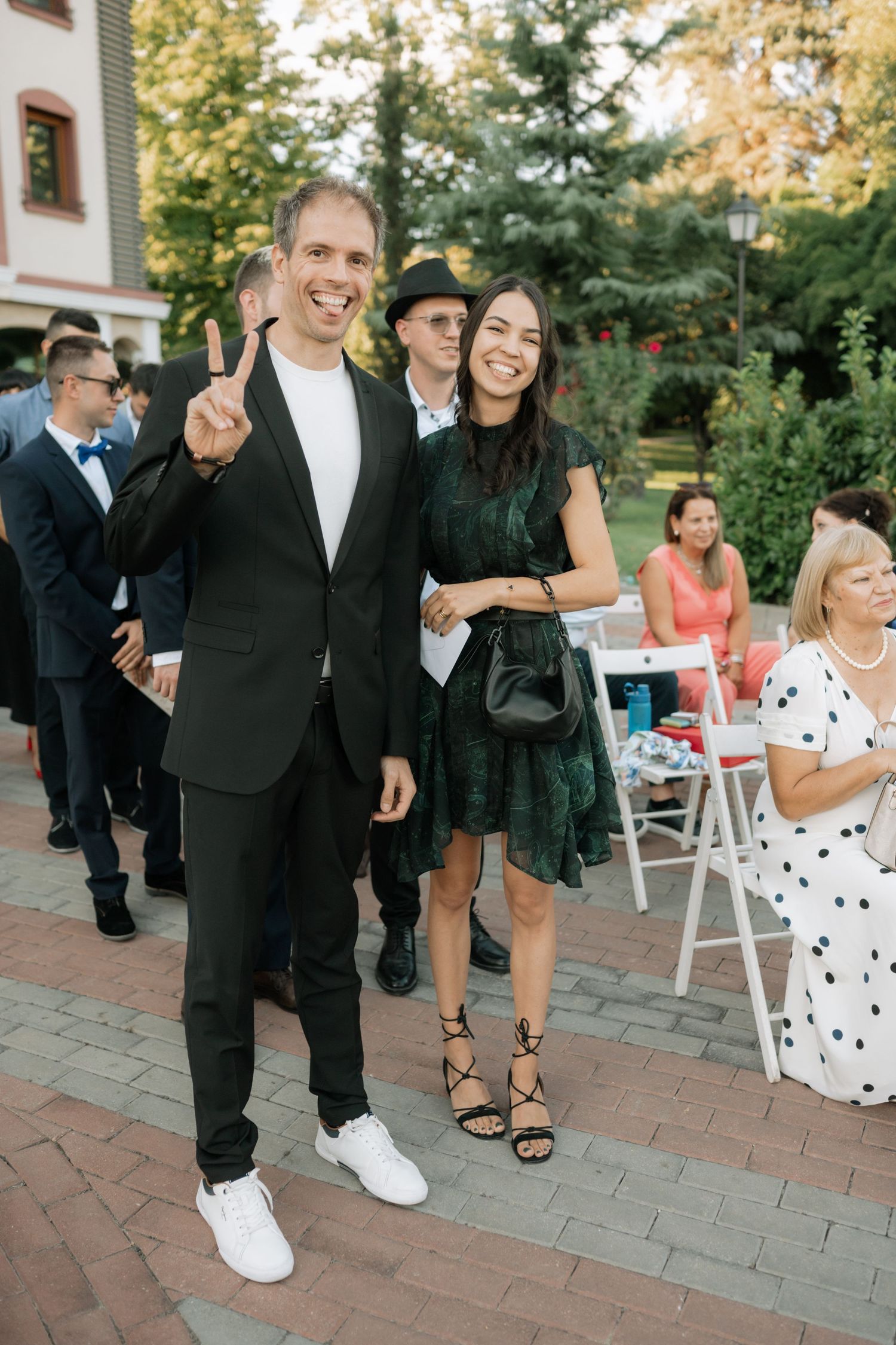 Wedding guests in formal attire smile and pose on a brick patio during an outdoor reception.