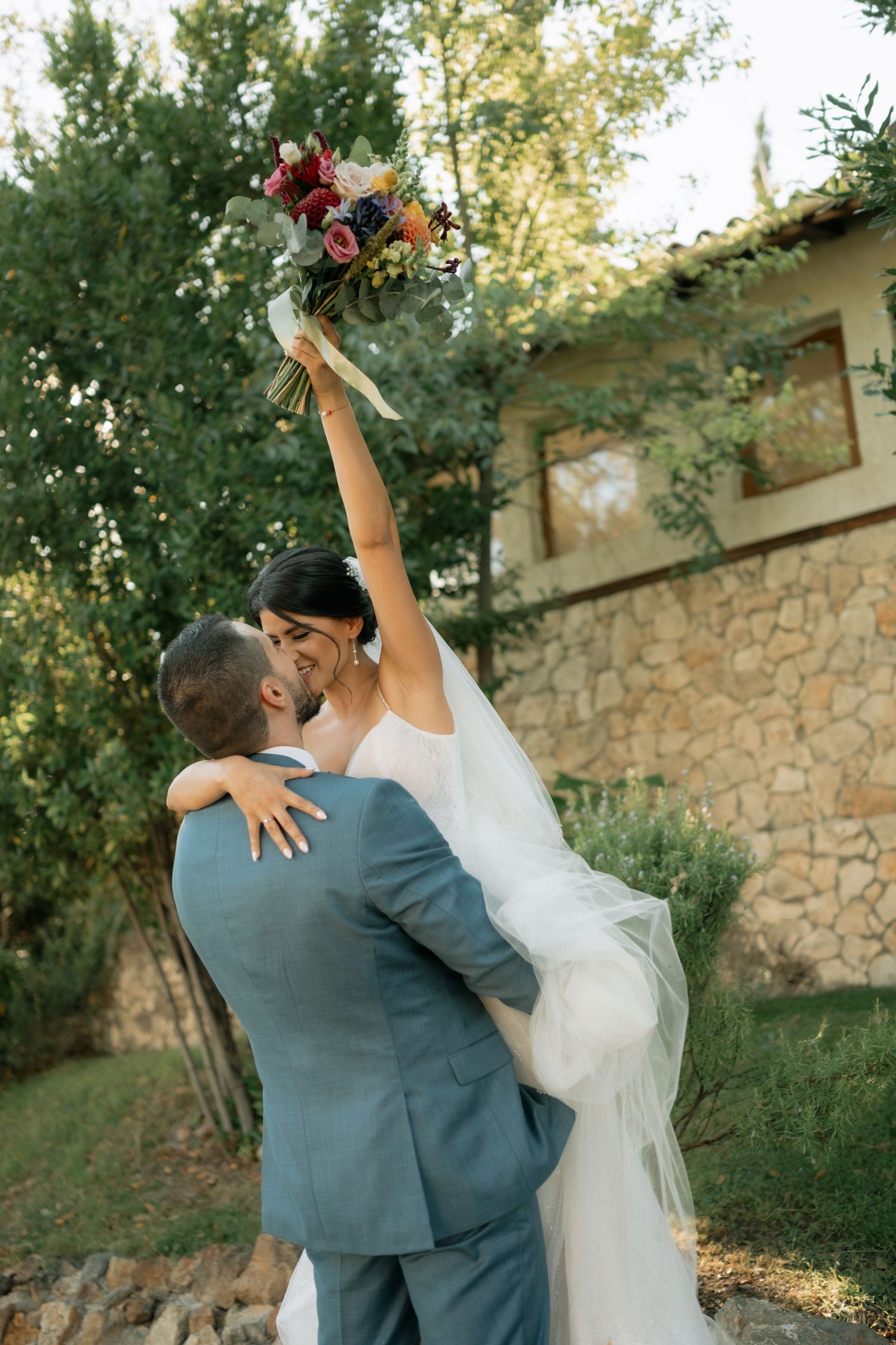 A couple in wedding attire dances and embraces in a garden setting with a stone wall backdrop during golden hour.