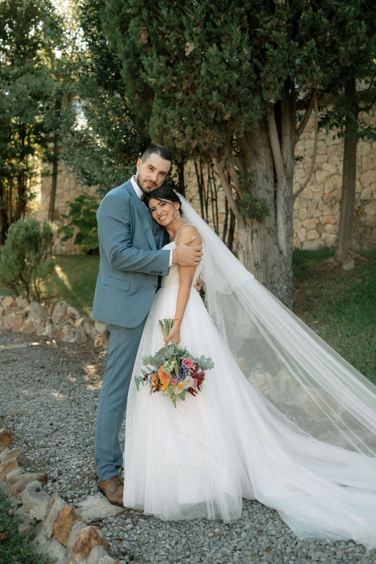 A romantic wedding photoshoot in a garden setting with a flowing white veil and gray suit.