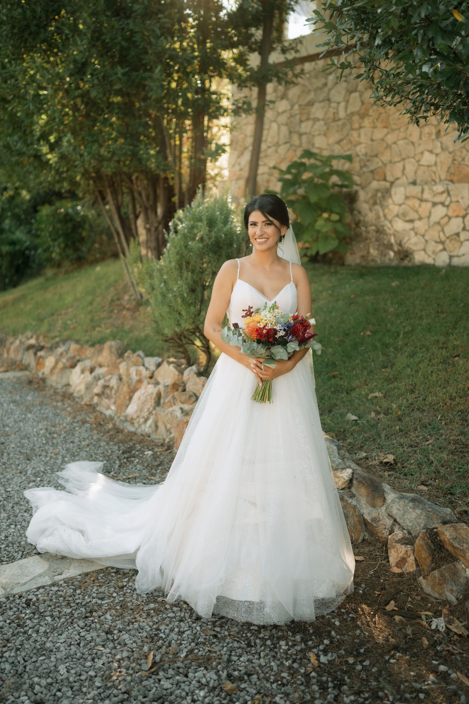 A bride in a flowing white ballgown poses gracefully against a rustic stone wall and natural outdoor setting.
