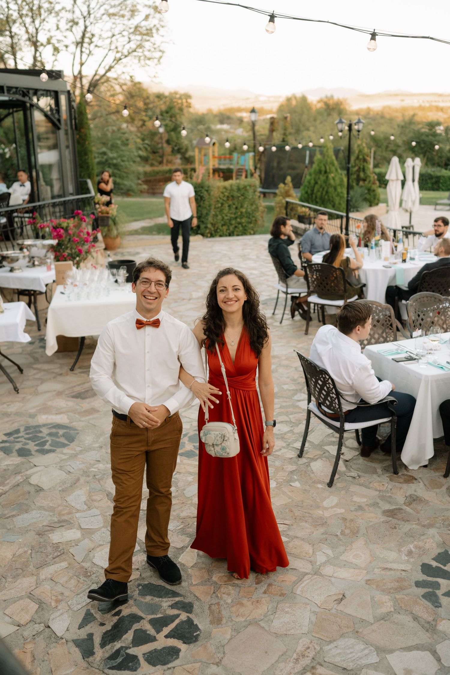 Outdoor wedding reception setup with string lights and stone patio featuring guests in formal attire at sunset.