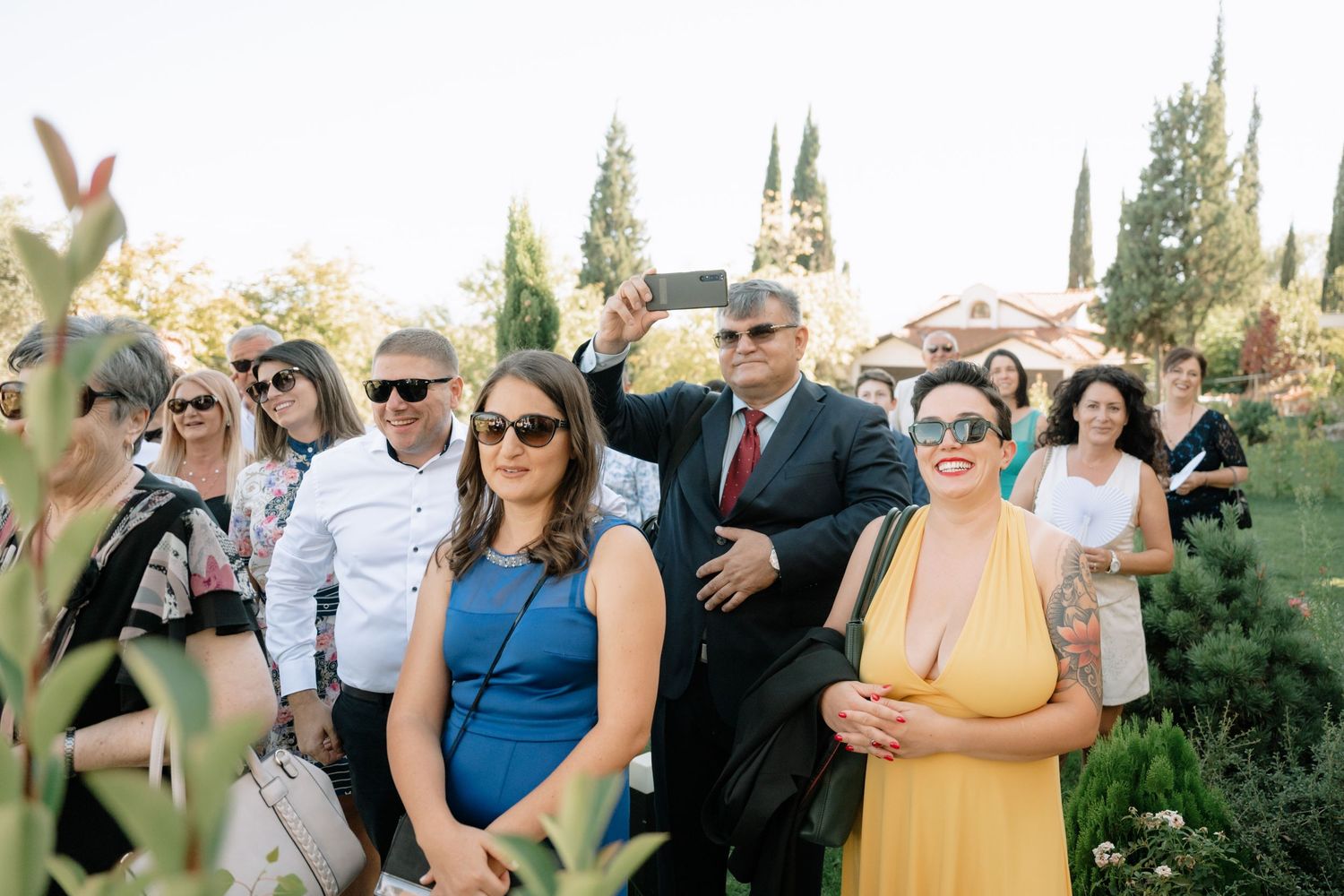Wedding guests in formal attire mingle and socialize outdoors during a sunny celebration.