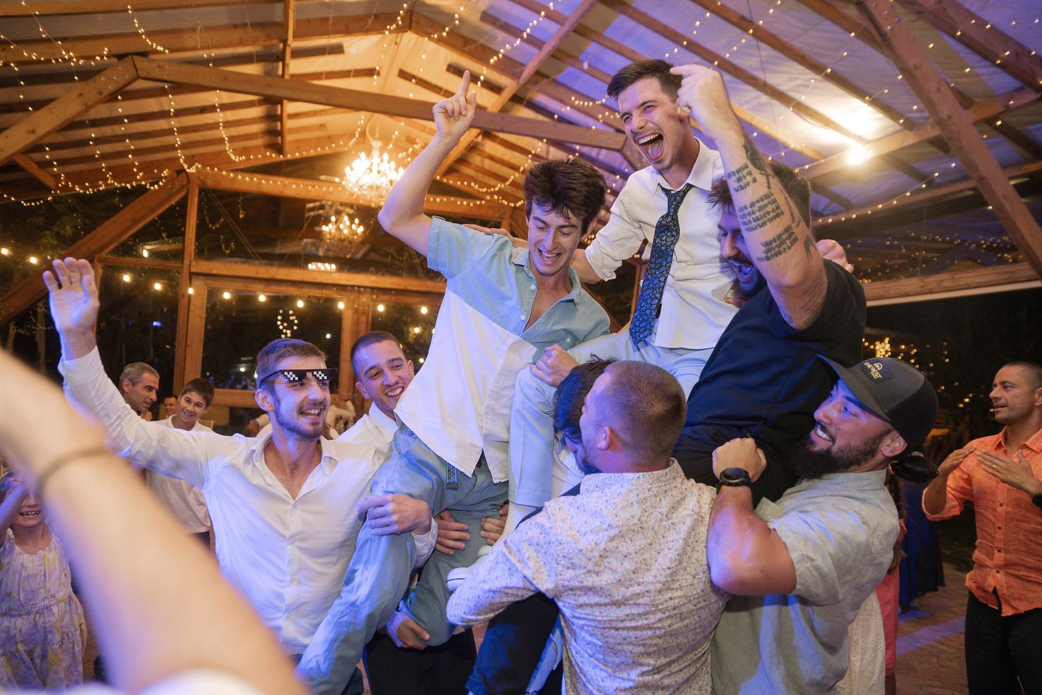Wedding guests dancing and celebrating together on the dance floor under colorful lighting at a reception venue.