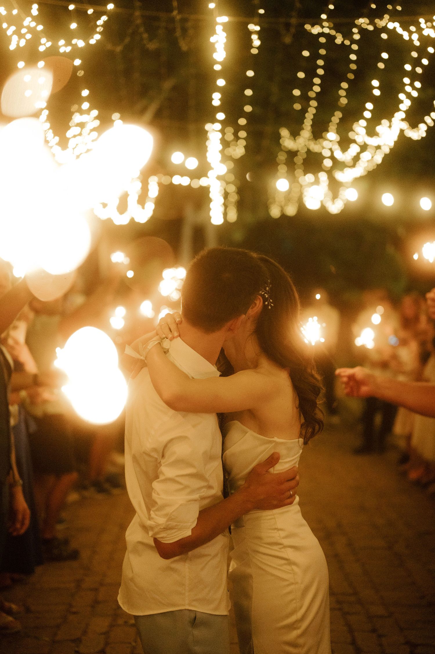 A romantic couple embraces under twinkling string lights at an evening outdoor celebration.