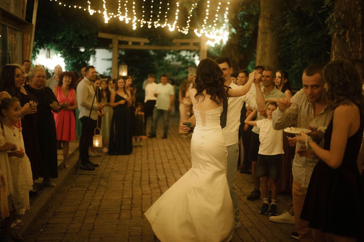 A bride in a white wedding dress dances under twinkling string lights at an outdoor evening reception.