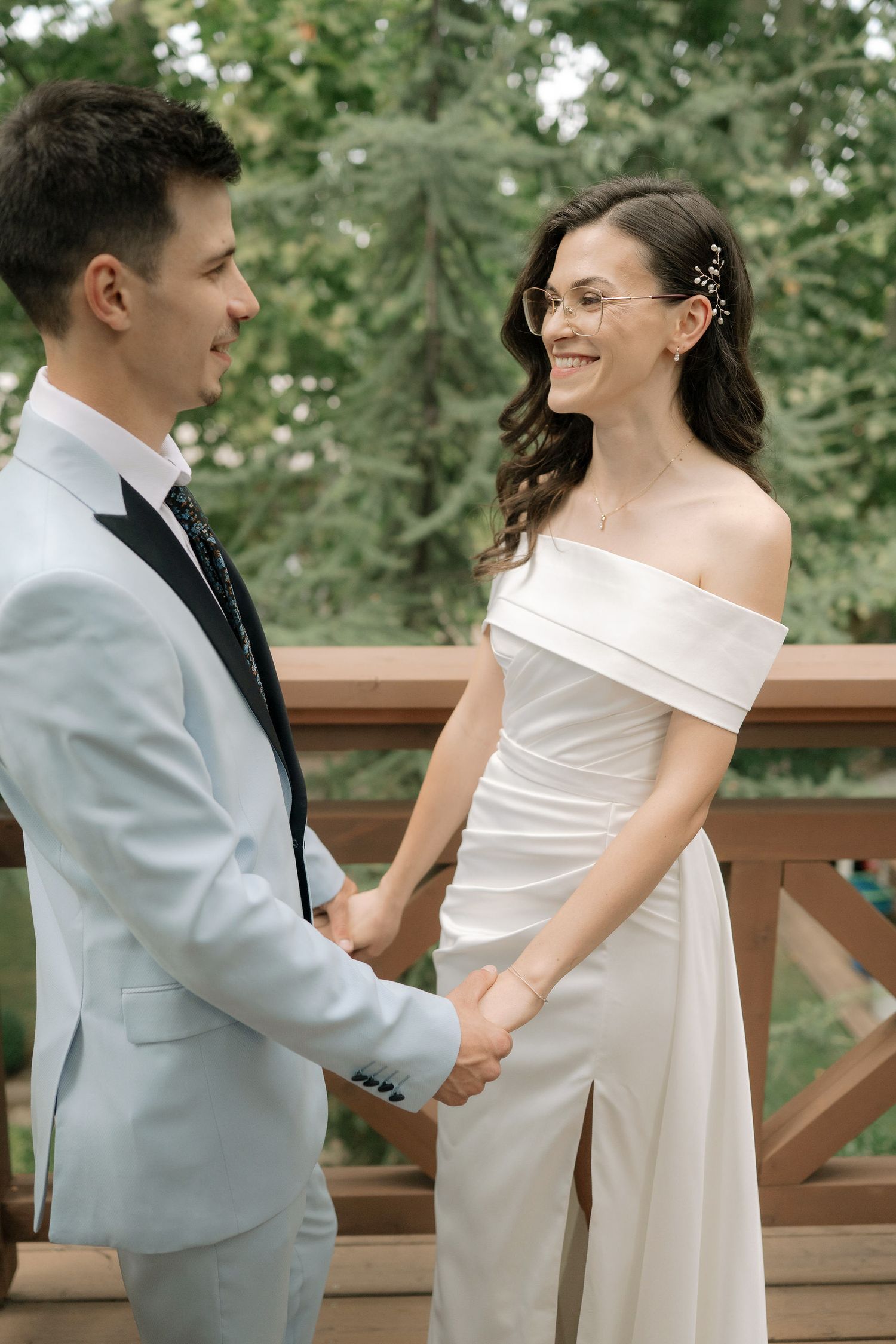 A couple in wedding attire share a joyful moment on a wooden deck surrounded by green trees.
