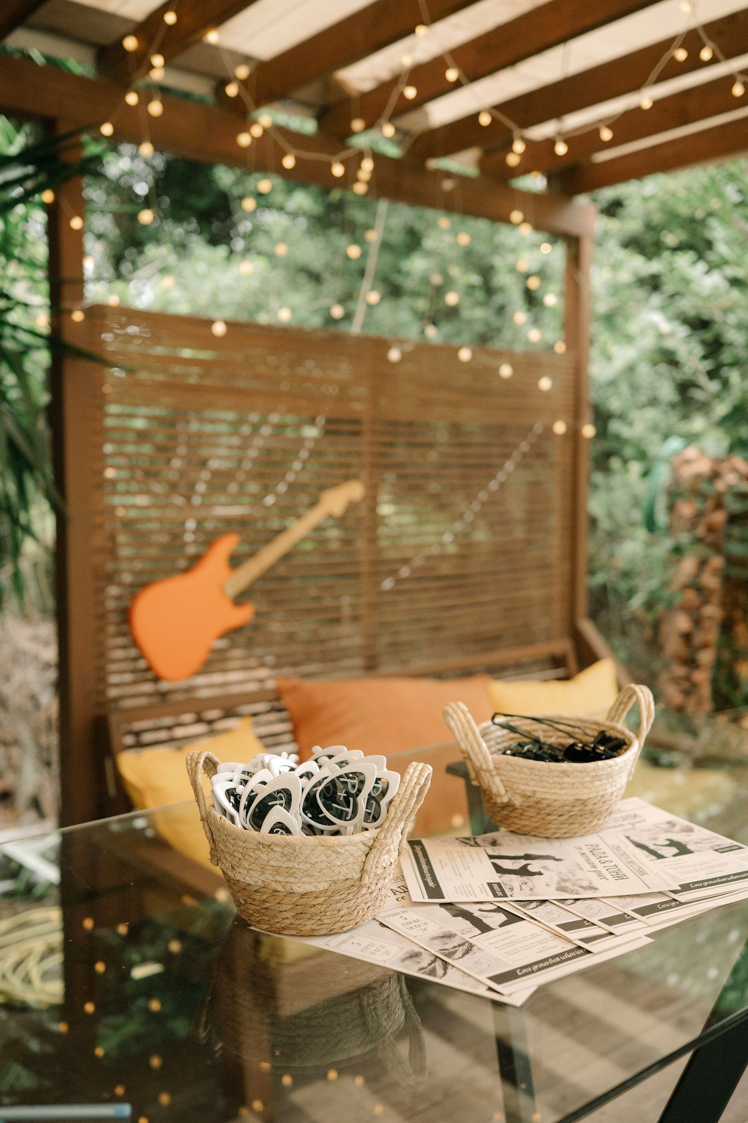 Rustic outdoor patio setup with string lights, guitar decor, and woven baskets on a glass table in a garden setting.