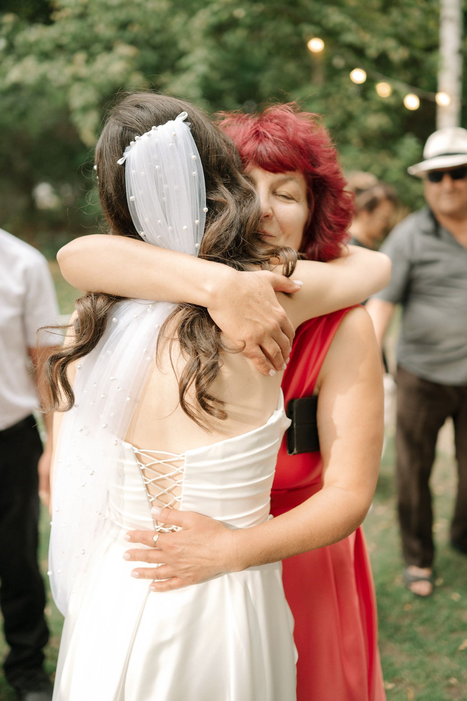 A bride in a white lace-up dress embraces someone in a red dress at an outdoor wedding celebration.