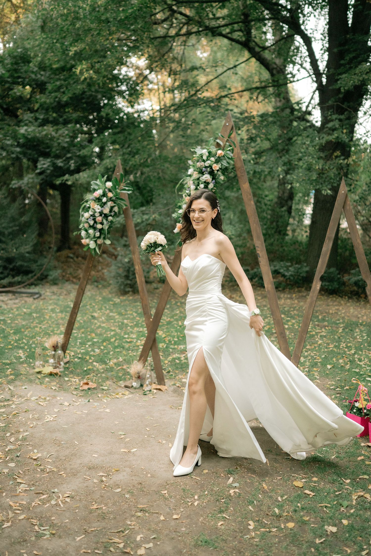 A bride in an elegant white gown dances joyfully through a scenic outdoor garden path surrounded by trees.