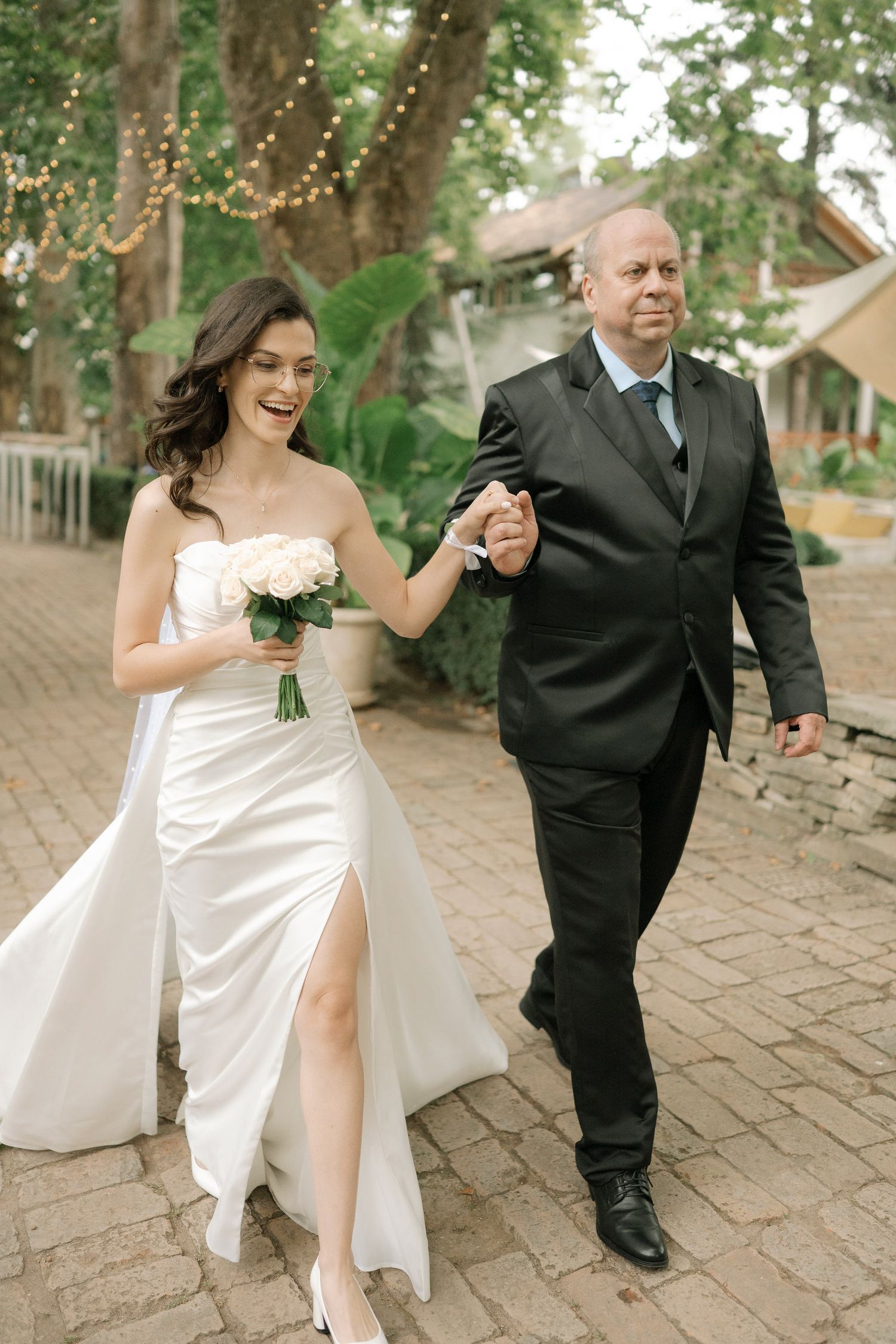 A bride and groom dance together on a brick pathway surrounded by lush green trees.
