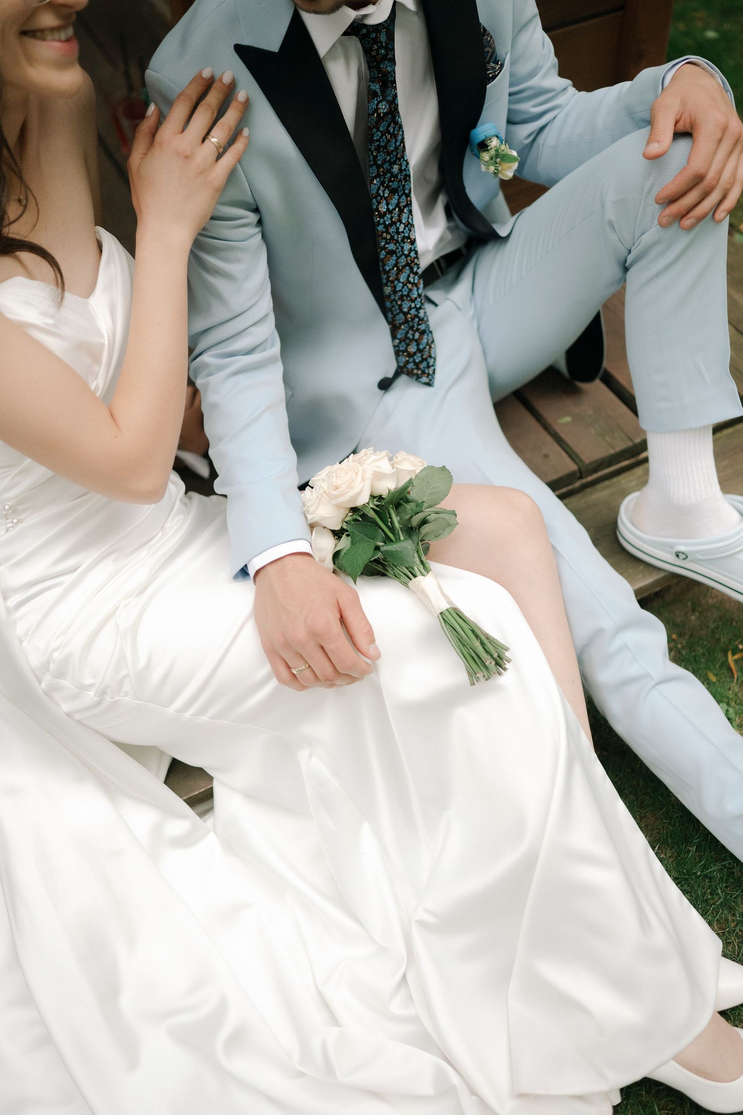 A couple sits together outdoors at a wedding, with a light blue suit, white dress and small bouquet of roses.