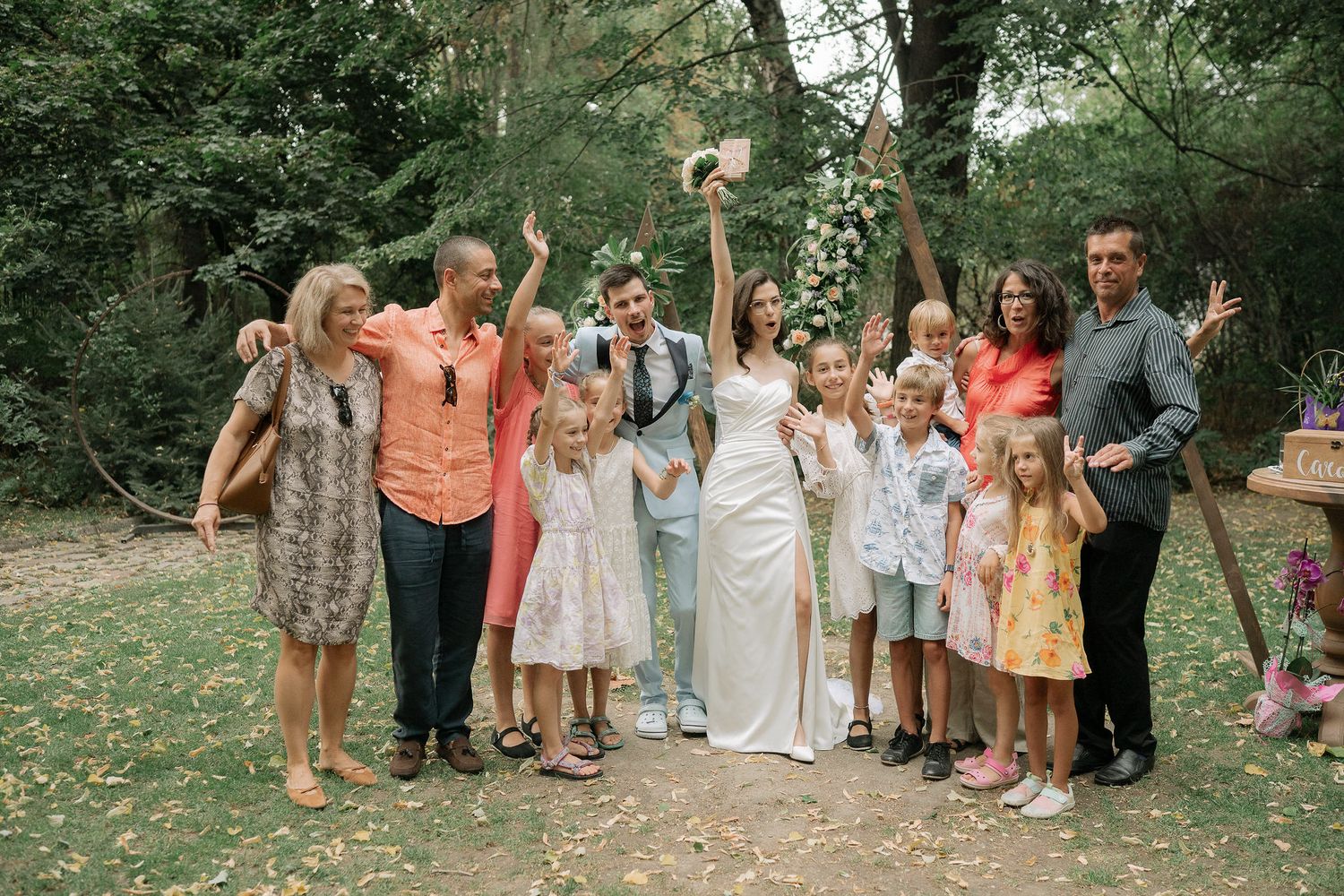 A group of wedding guests in coral and pastel dresses pose together outdoors in a garden setting.