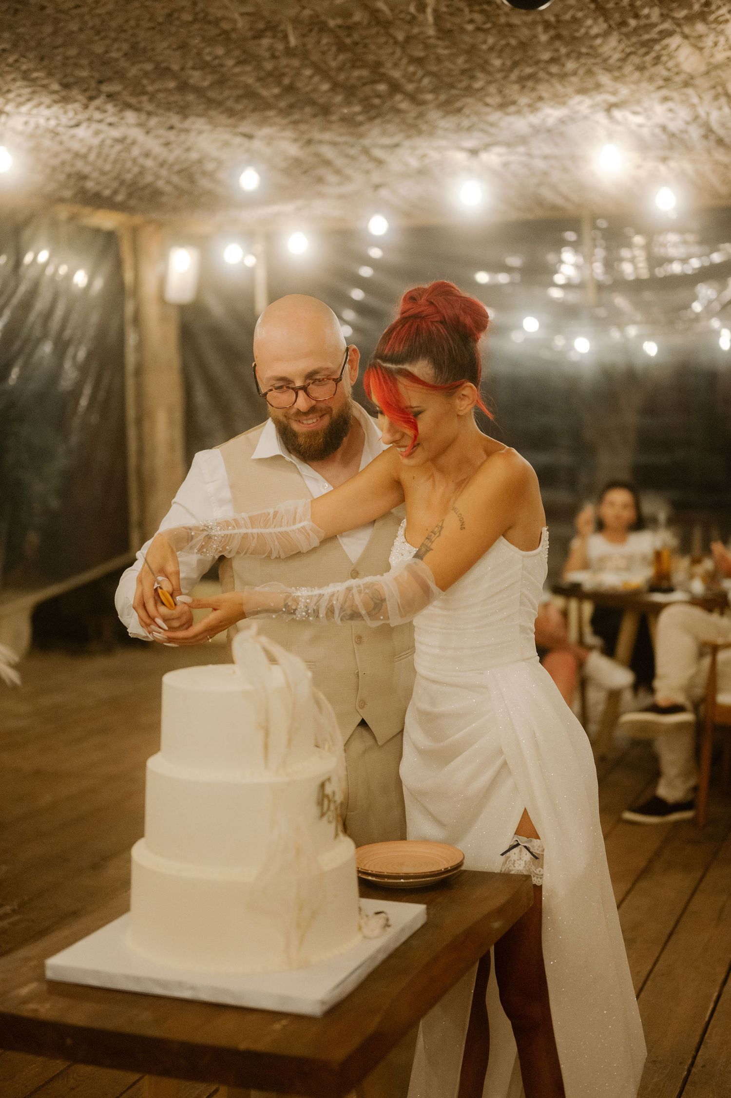 Couple playfully cutting their white wedding cake together at a reception with string lights overhead.