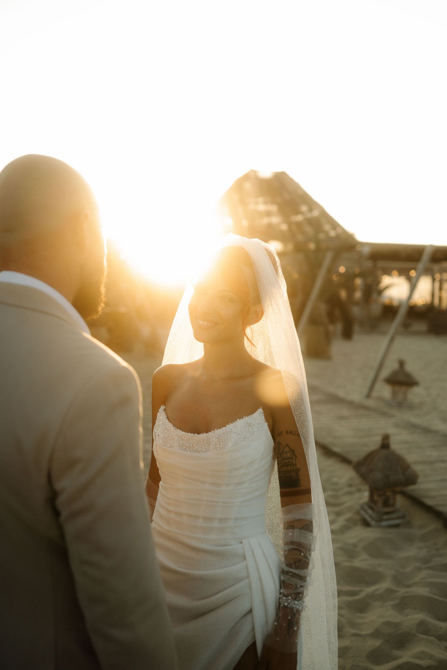 A couple shares an intimate moment on a beach at sunset with palm umbrellas in the background.