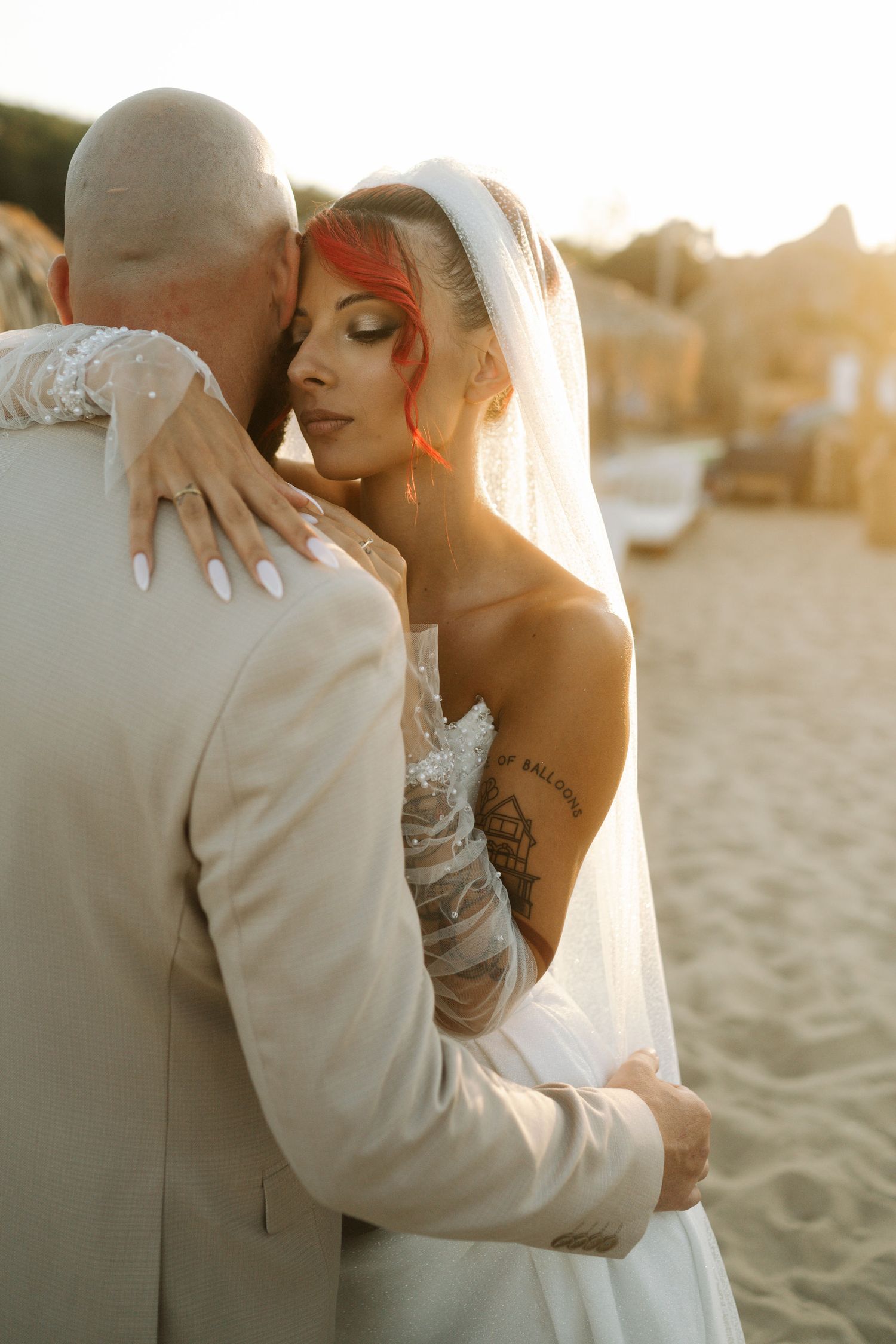 Romantic sunset beach wedding photo showing couple embracing in white attire with veil flowing in golden light.