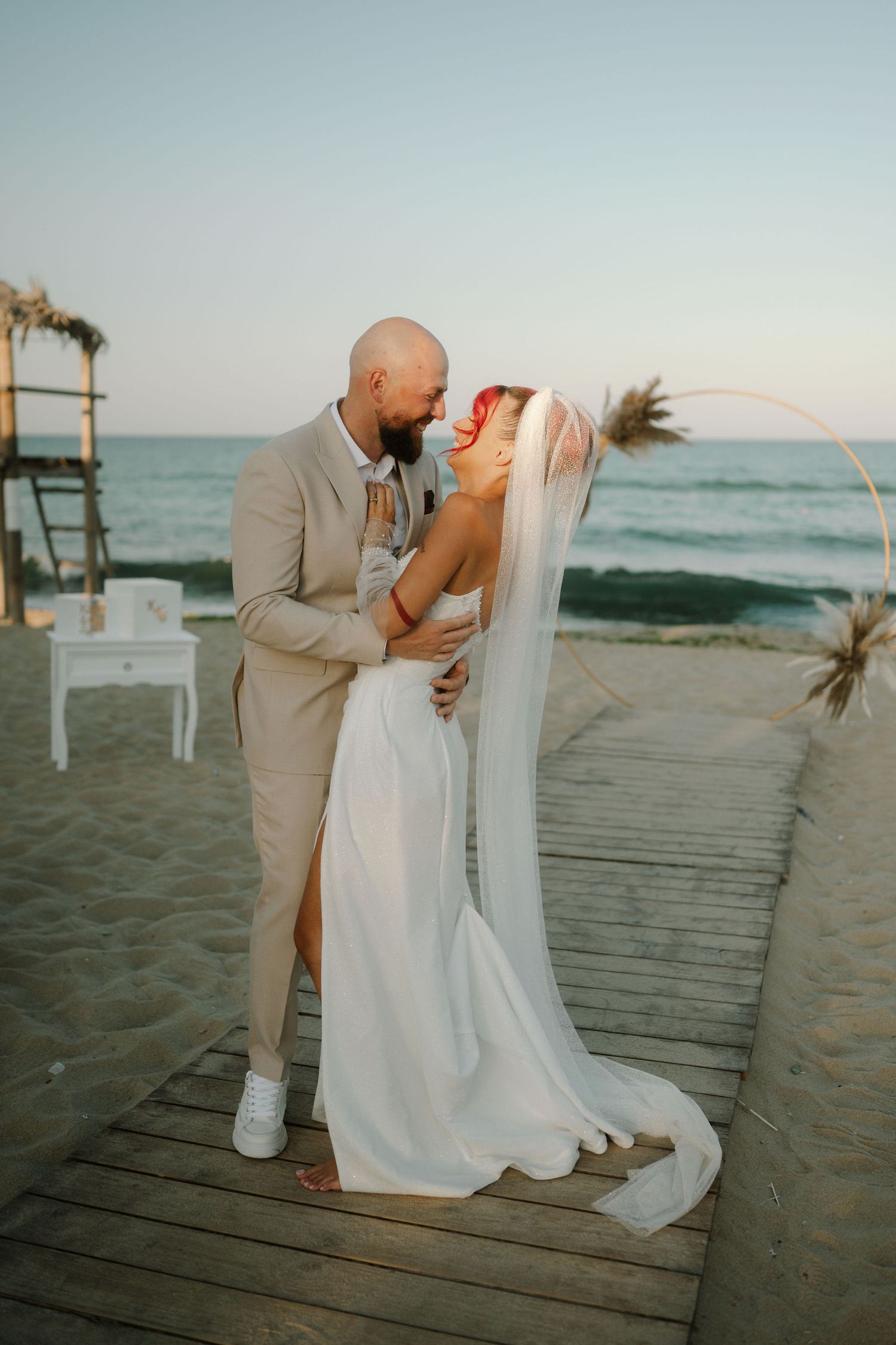 A romantic beach wedding couple shares intimate moments on a wooden boardwalk at sunset.