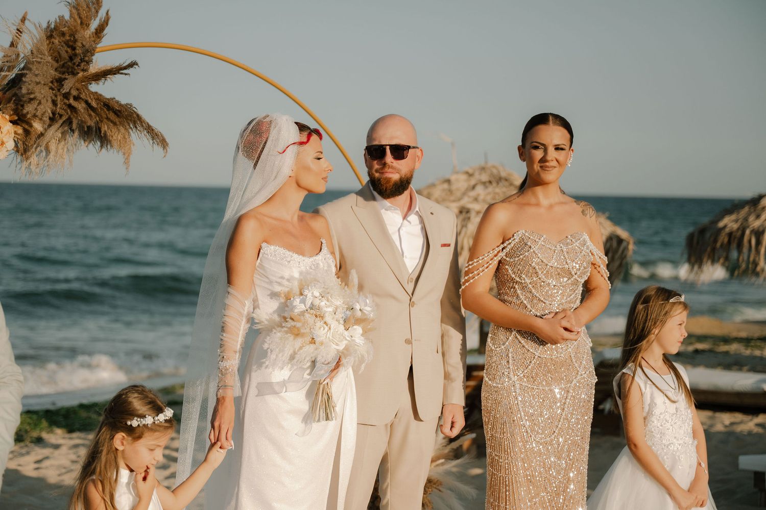 A beachside wedding party poses together at sunset with palm fronds and ocean waves in the background.