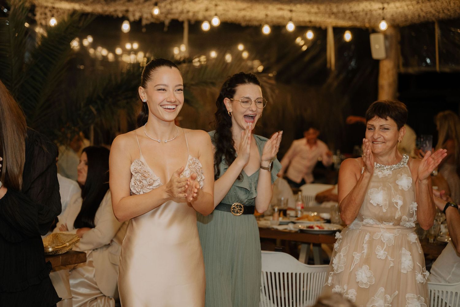Wedding guests clapping and celebrating at an evening reception under string lights and decorations.