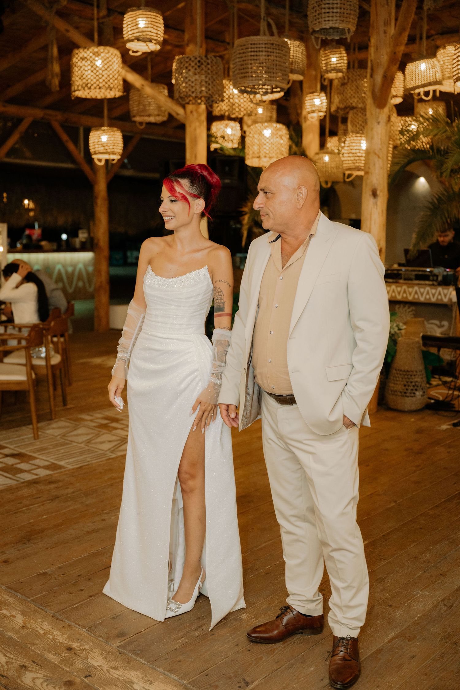A couple in matching white and beige outfits dances together under warm lighting at a rustic barn venue.