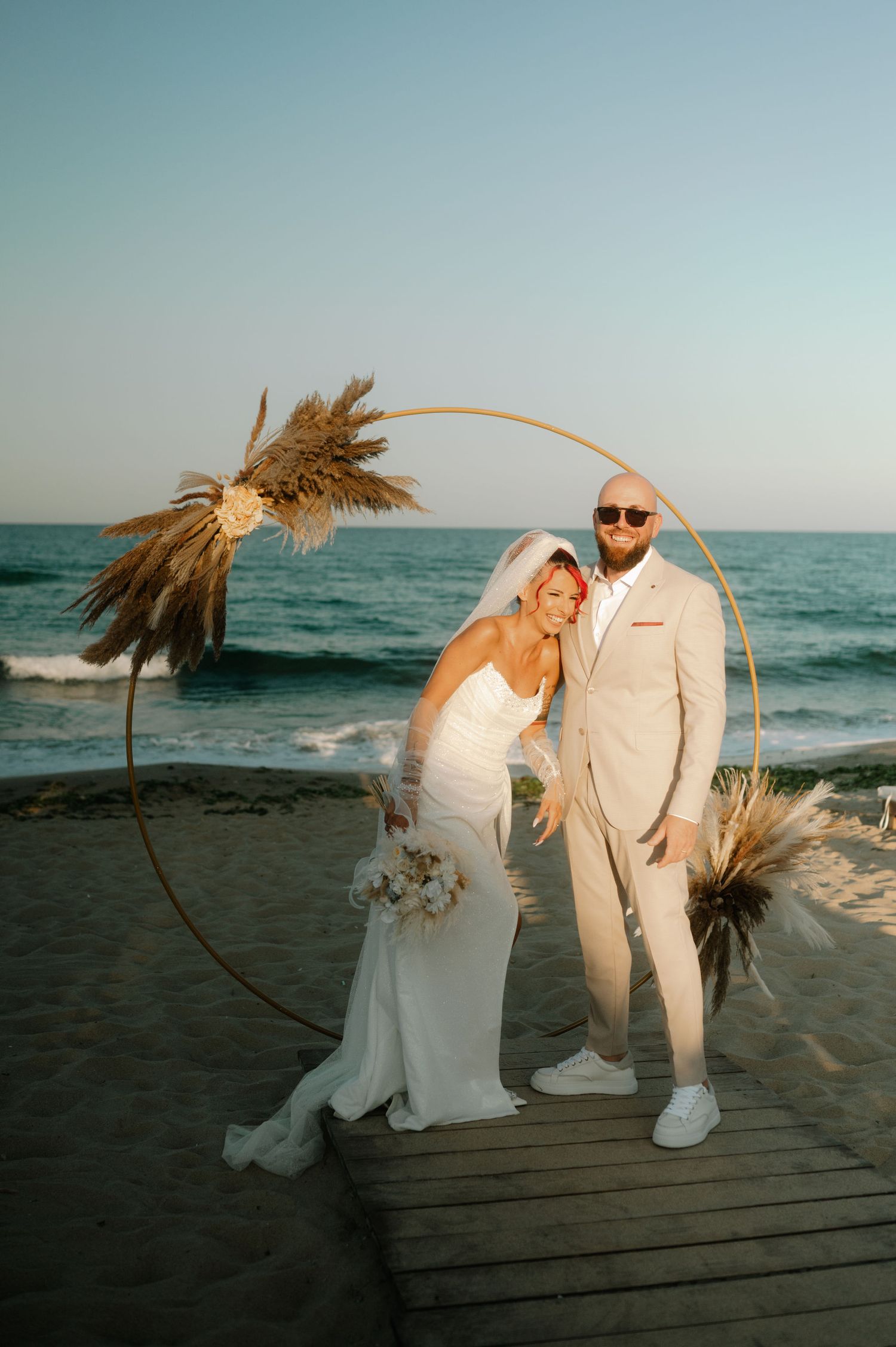A couple in wedding attire poses playfully on a wooden boardwalk at sunset by the beach with dried pampas grass decor.