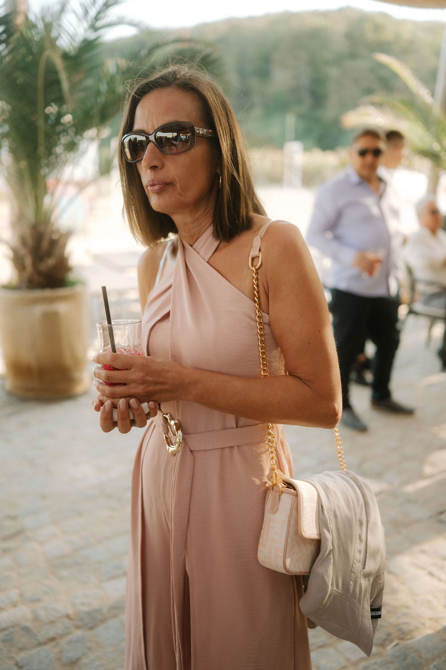 Elegant woman in blush pink one-shoulder dress holding drink at outdoor venue with palm trees in background.