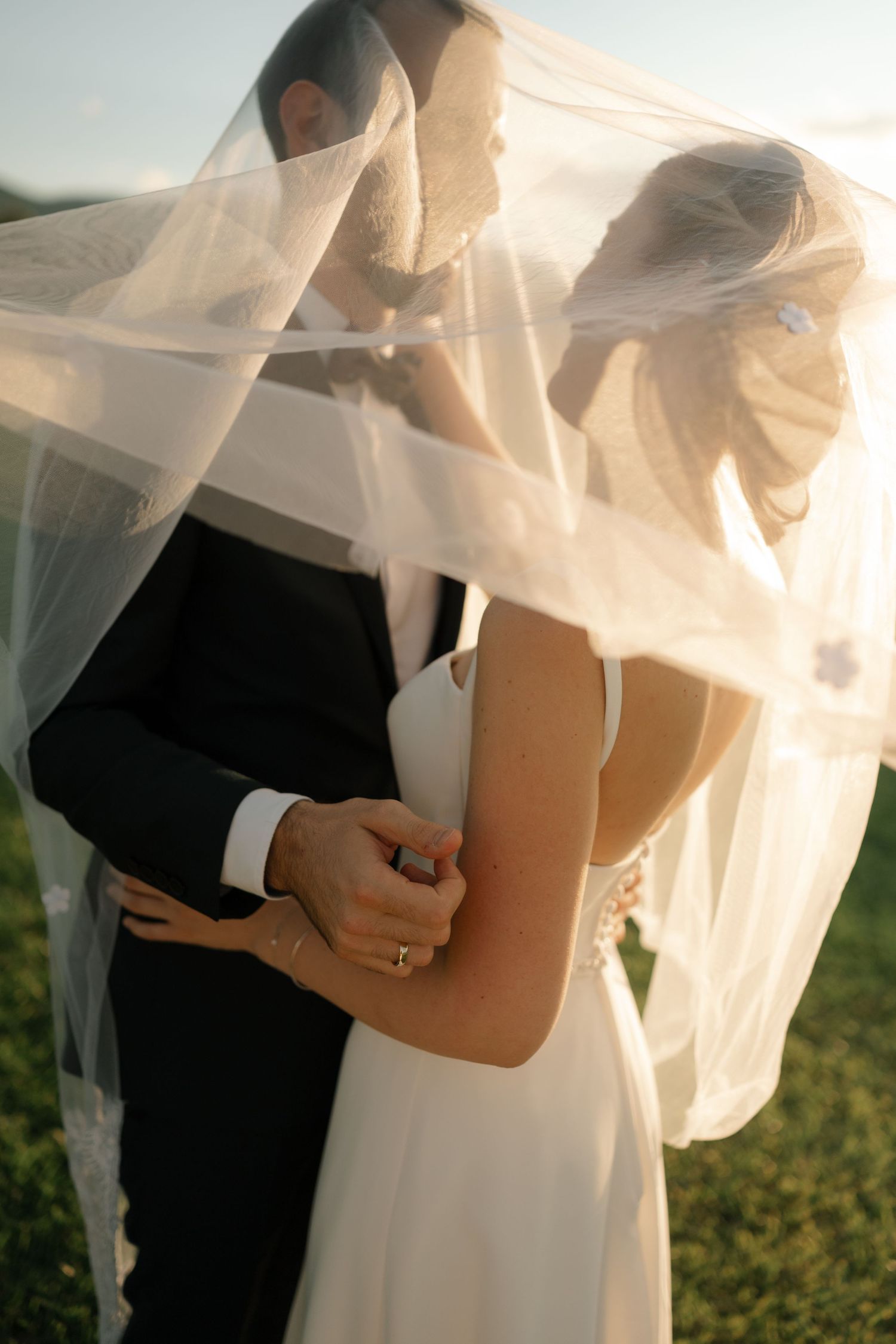 Romantic wedding couple embraces under flowing white veil during outdoor sunset ceremony.