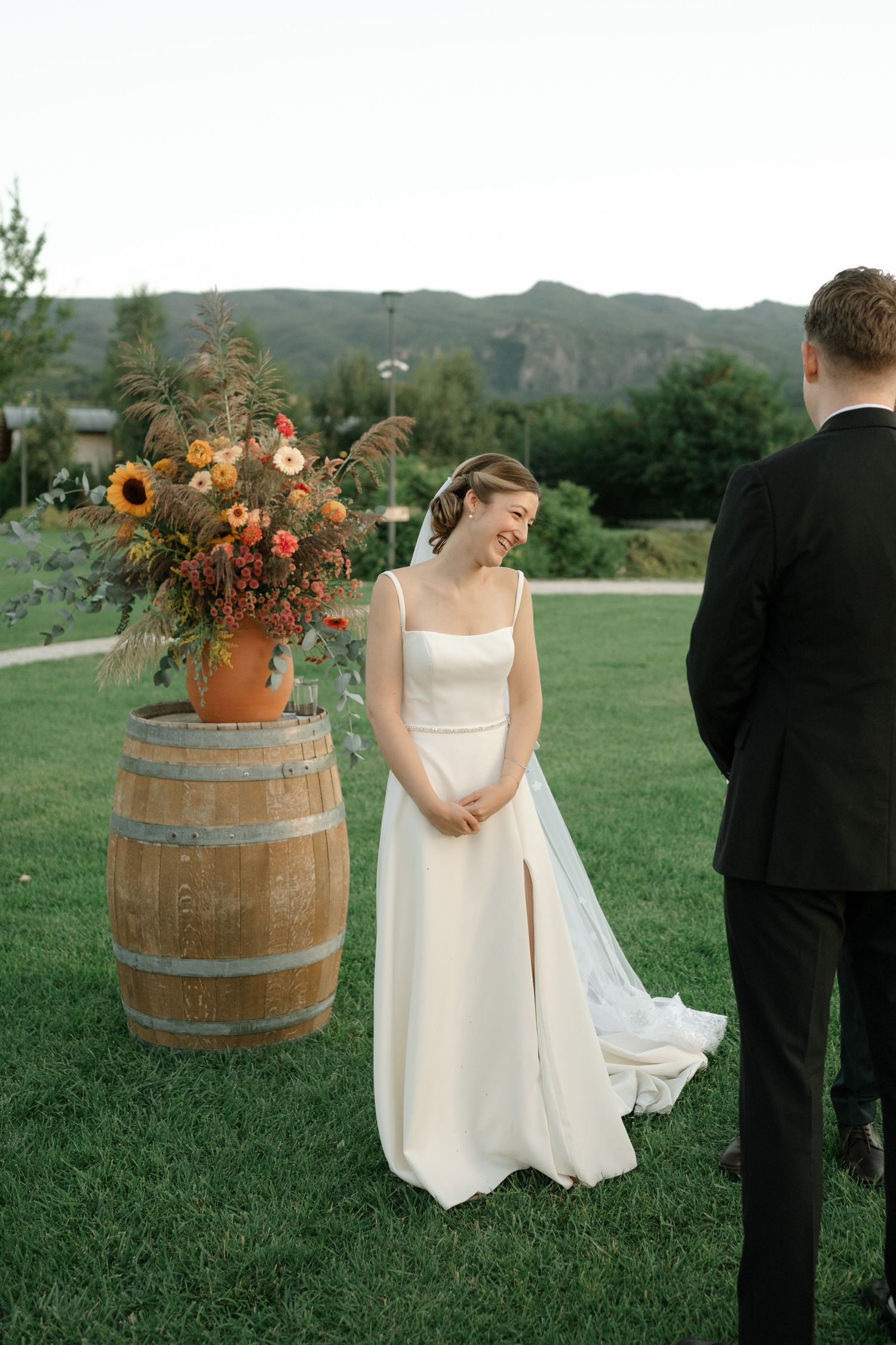 A wedding couple stands in a grassy field with rustic wine barrels and floral arrangements creating a romantic outdoor setting.