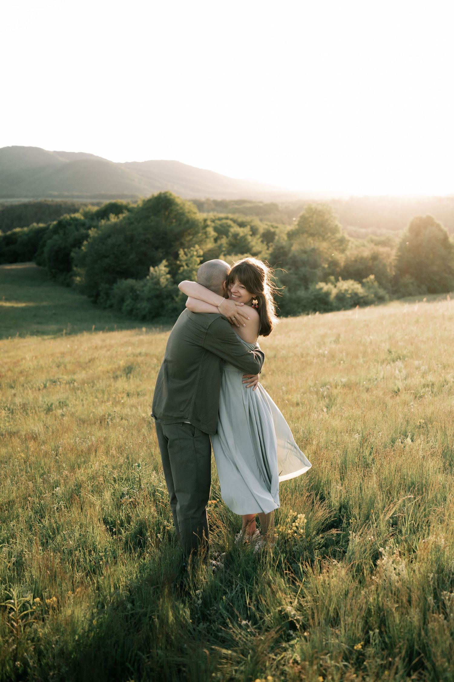 Couple embracing in a sunlit meadow with rolling green hills and trees in the background at golden hour.