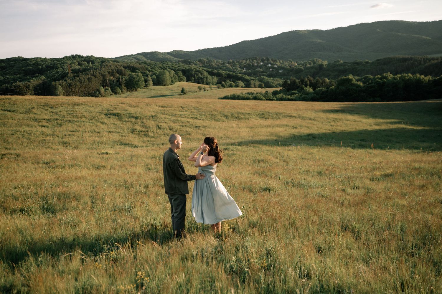 Couple holding hands in a lush green meadow with rolling hills and forest backdrop at golden hour.