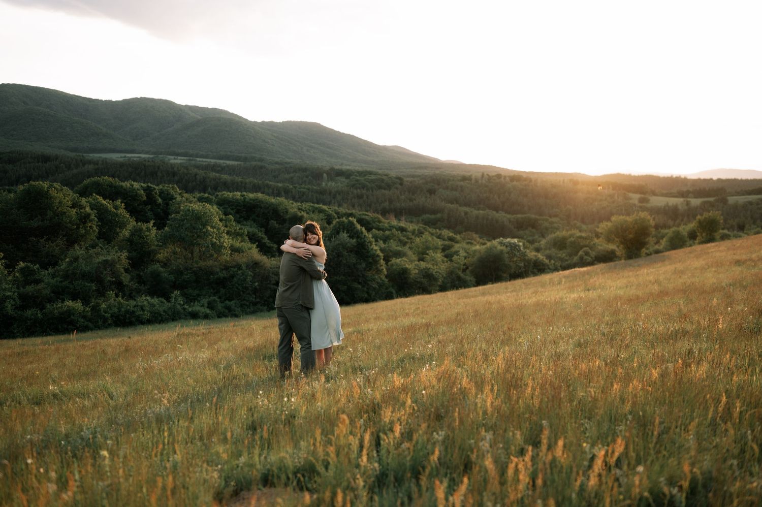 Couple embracing in a golden sunlit meadow with lush green hills and forest in the background at sunset.