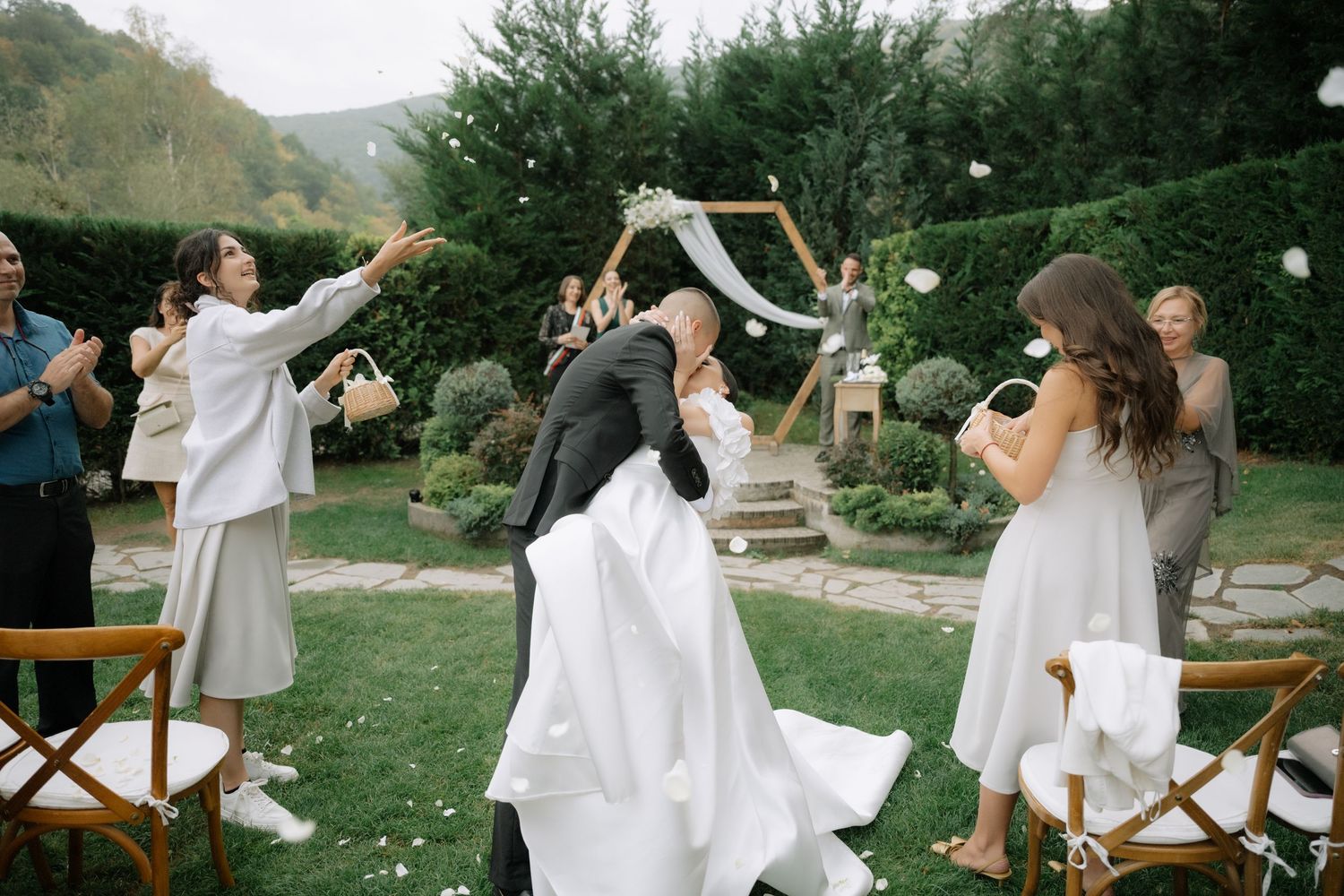 Joyful celebration at an outdoor garden wedding with guests throwing flower petals in the air.