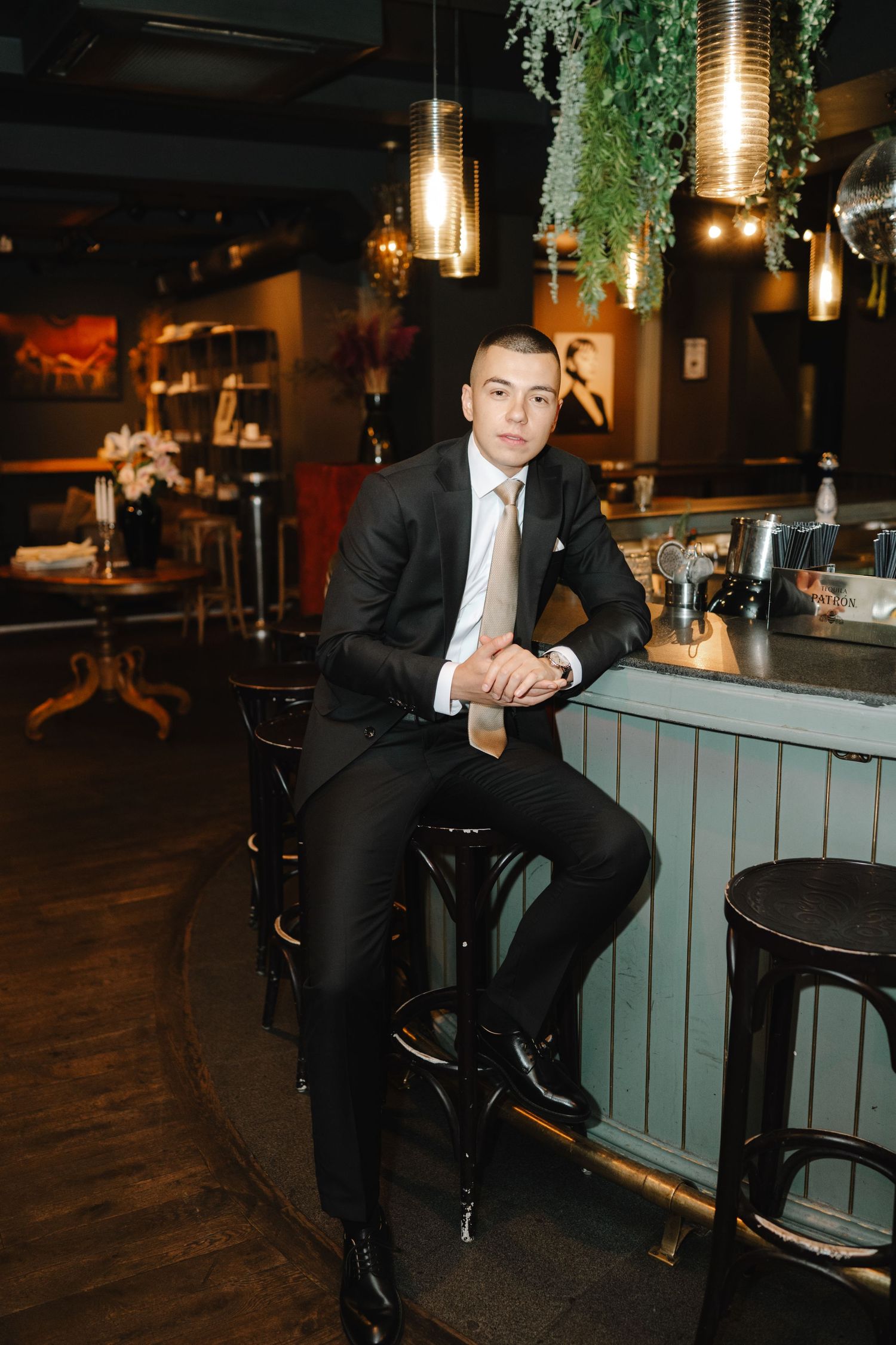A person in a black suit sits at a bar counter with hanging pendant lights and greenery decorations in a dark restaurant setting.