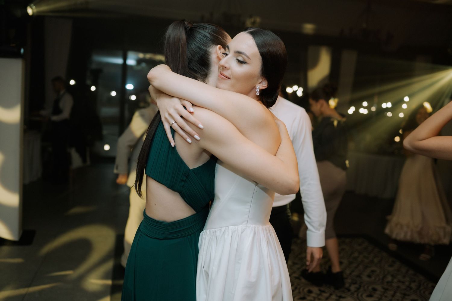 Two people embrace in an emotional moment at an evening event, one wearing a white dress and one in emerald green.