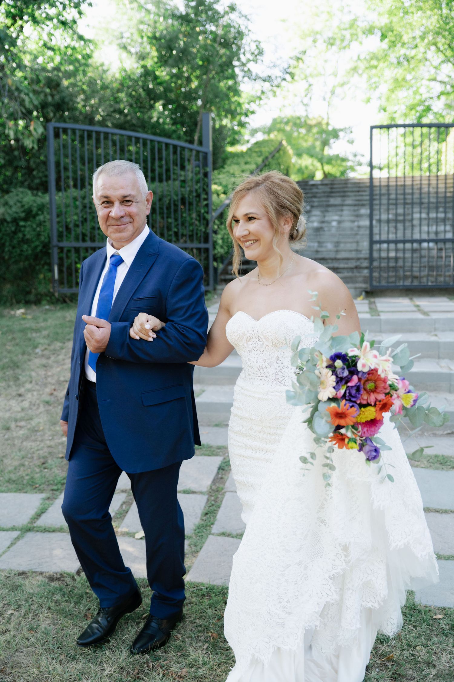 Bride in white dress poses with wedding party member in navy suit outdoors.