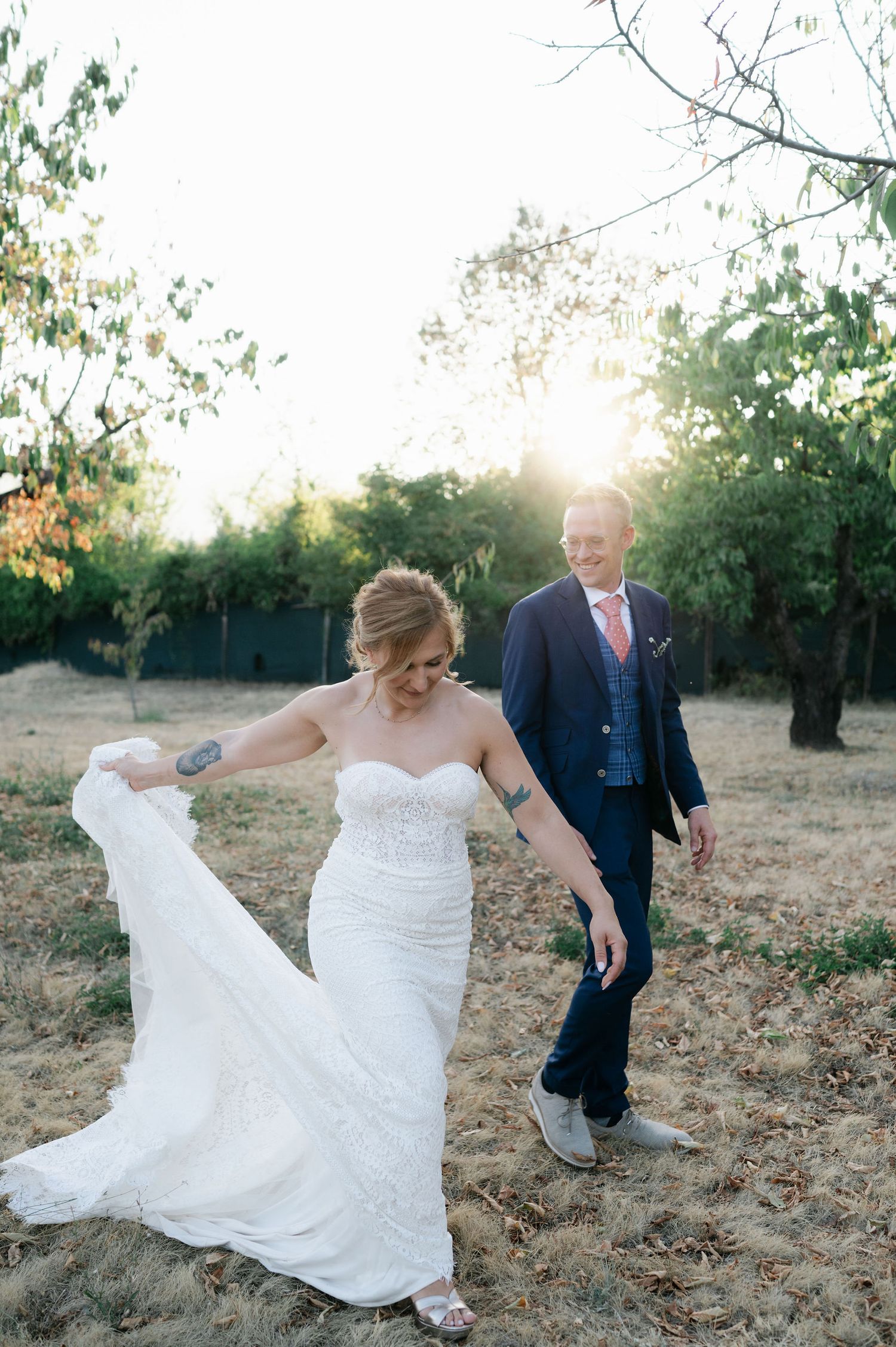 Bride in lace dress twirls her gown while groom watches on outdoor path at sunset.