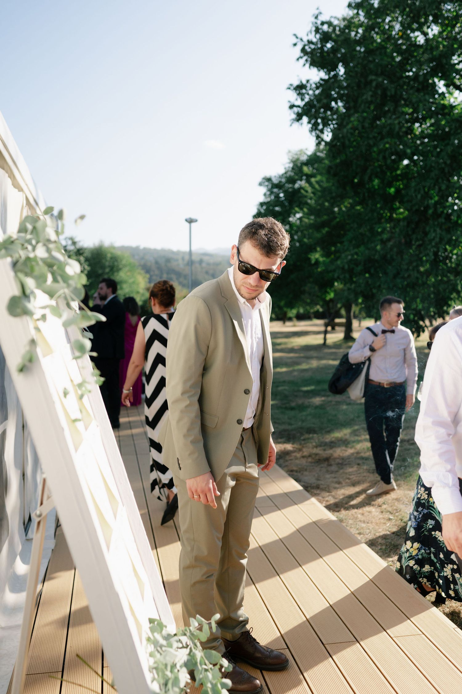 Wedding guests mingle on an outdoor wooden deck surrounded by greenery and white decorative elements.