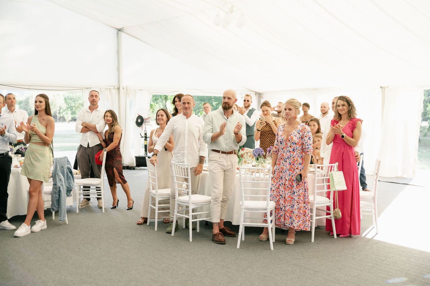 Wedding guests in formal attire standing and clapping during reception under white tent.
