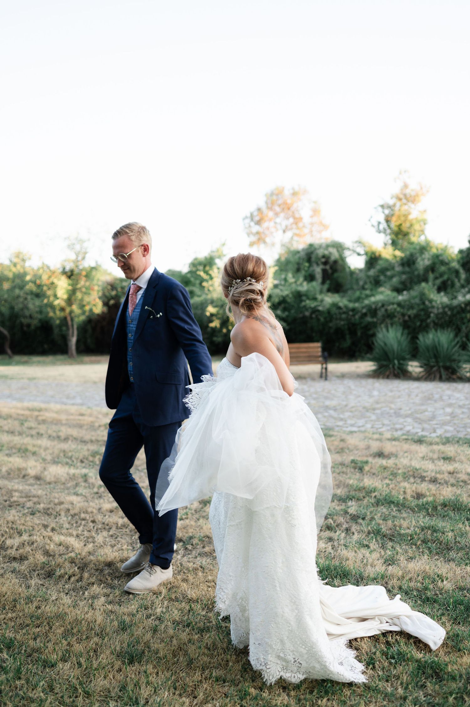 A couple in wedding attire walks through a grassy field at sunset.