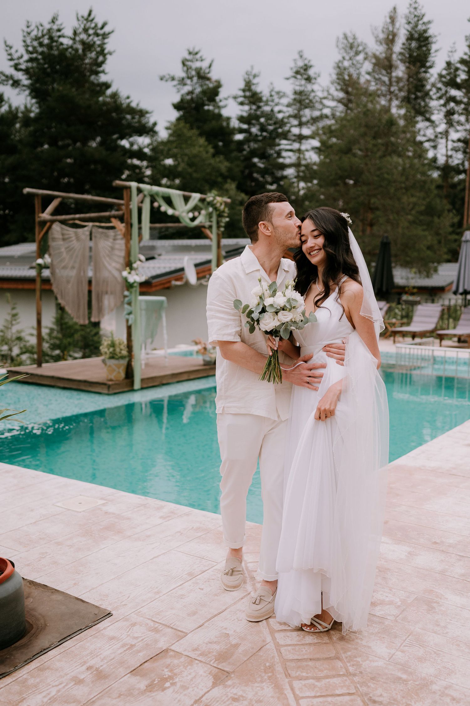 Romantic wedding photos captured by a turquoise pool with pine trees in the background.