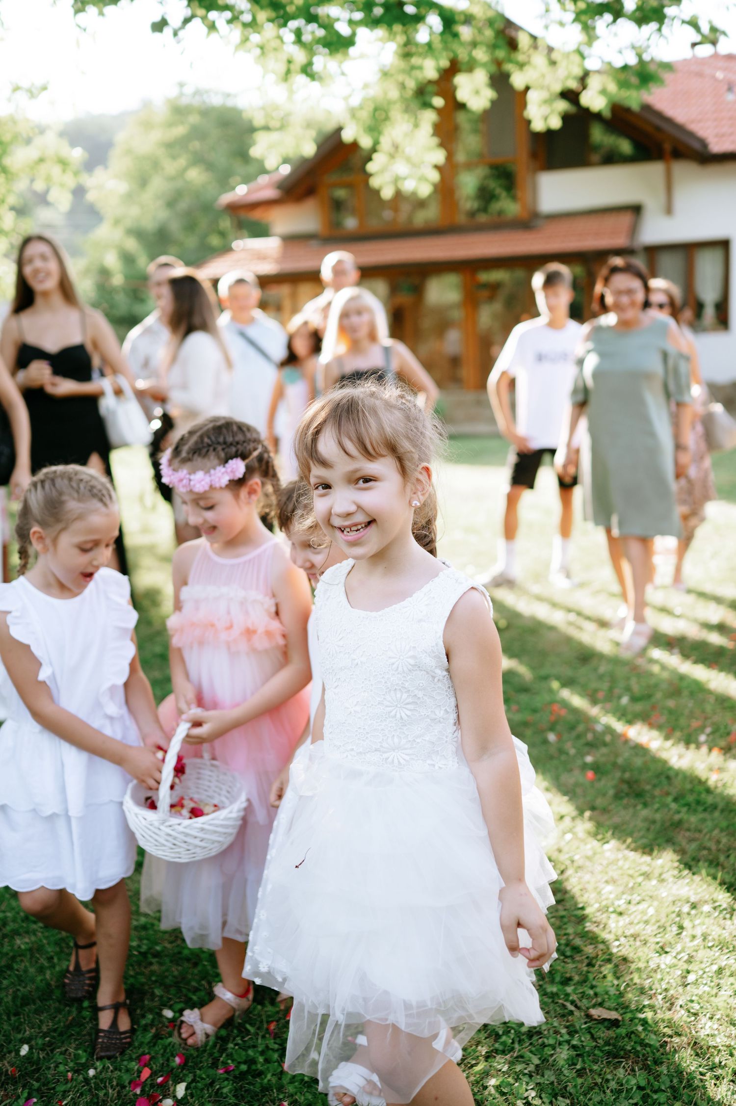 Children in white dresses carrying flower baskets at a sunny outdoor wedding celebration.