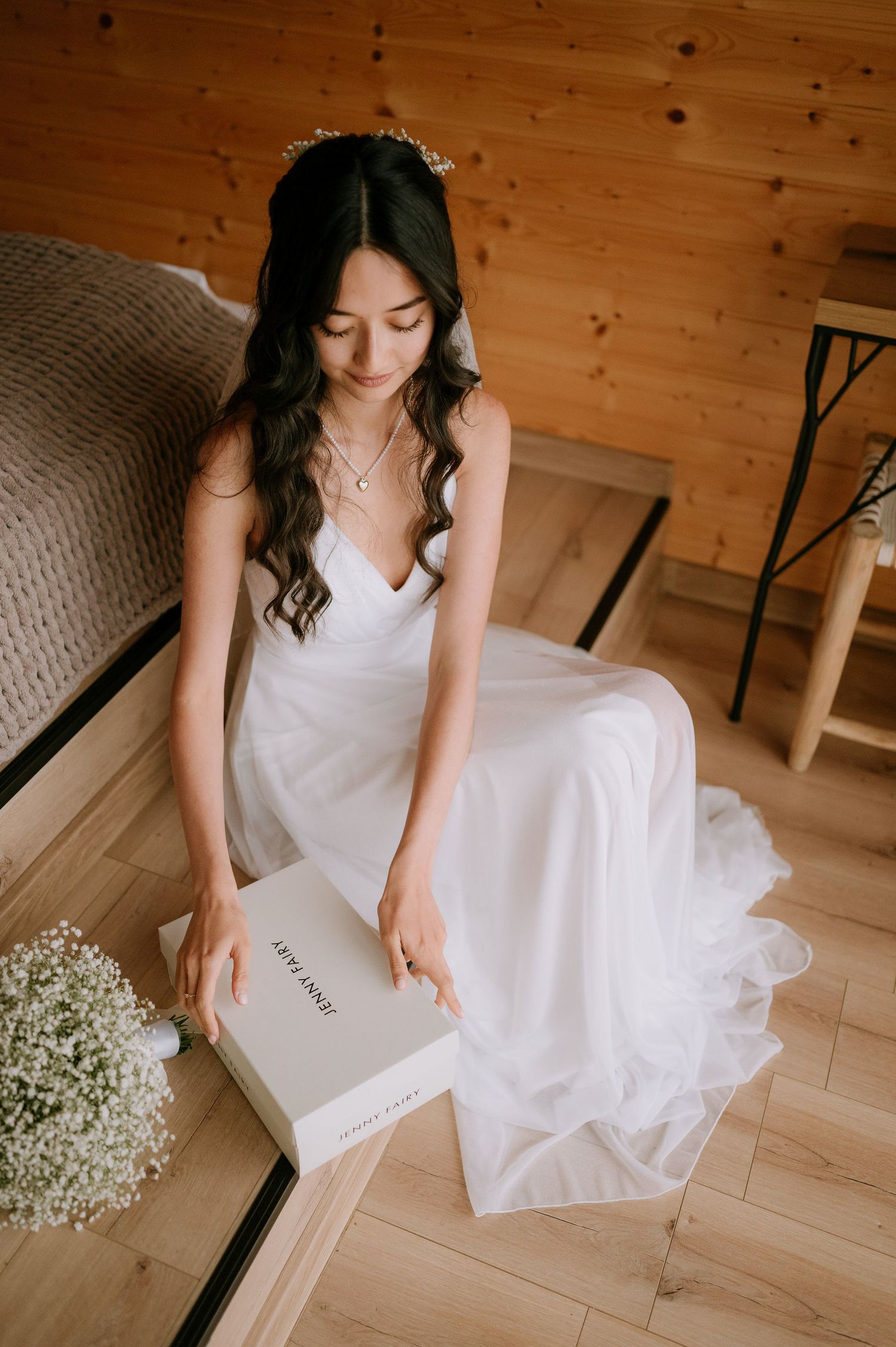 Bride in white dress sits with a book, surrounded by baby's breath flowers in a rustic setting.