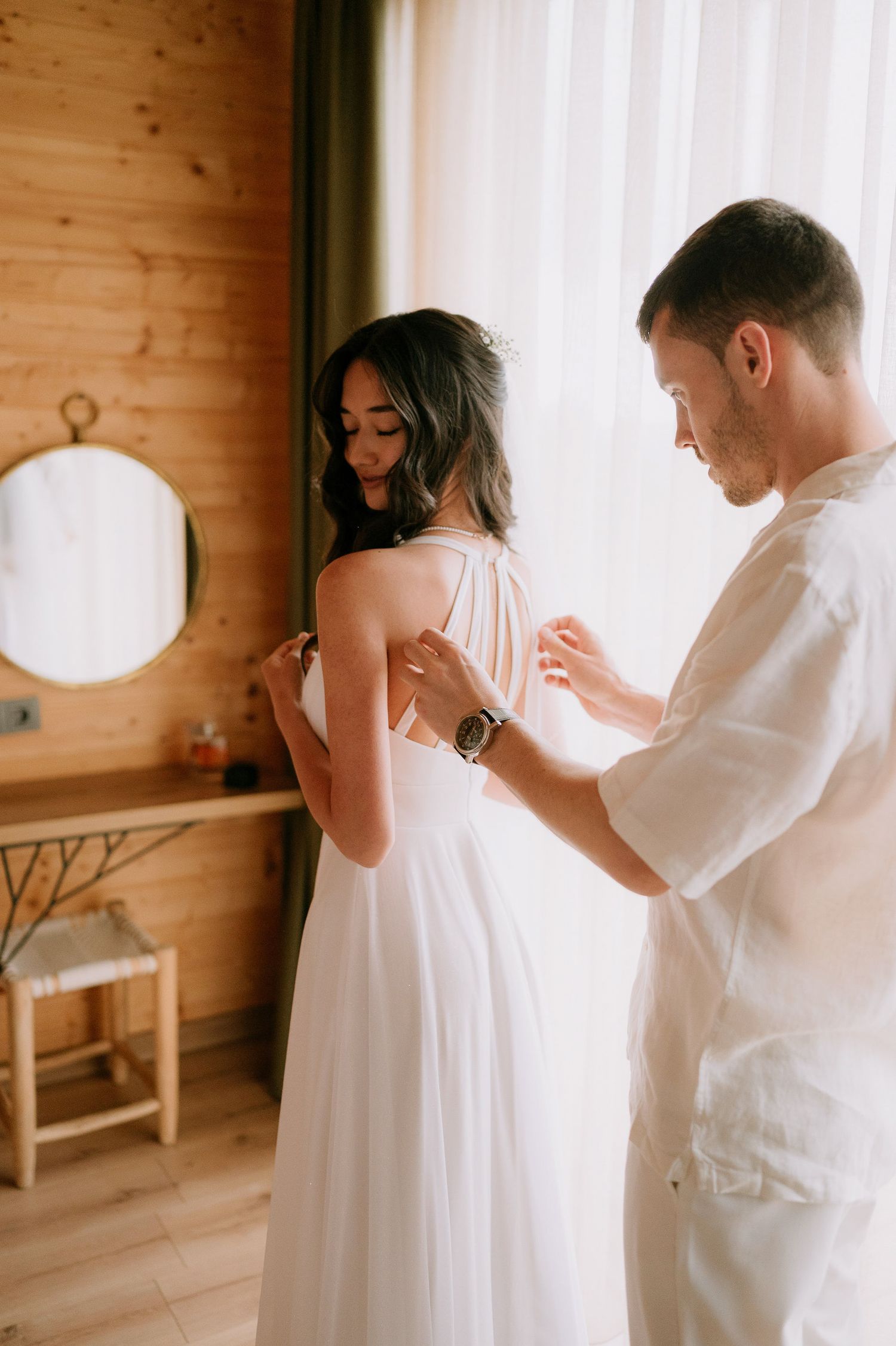 Intimate wedding preparation scene in a sunlit room with wooden walls and a round mirror.