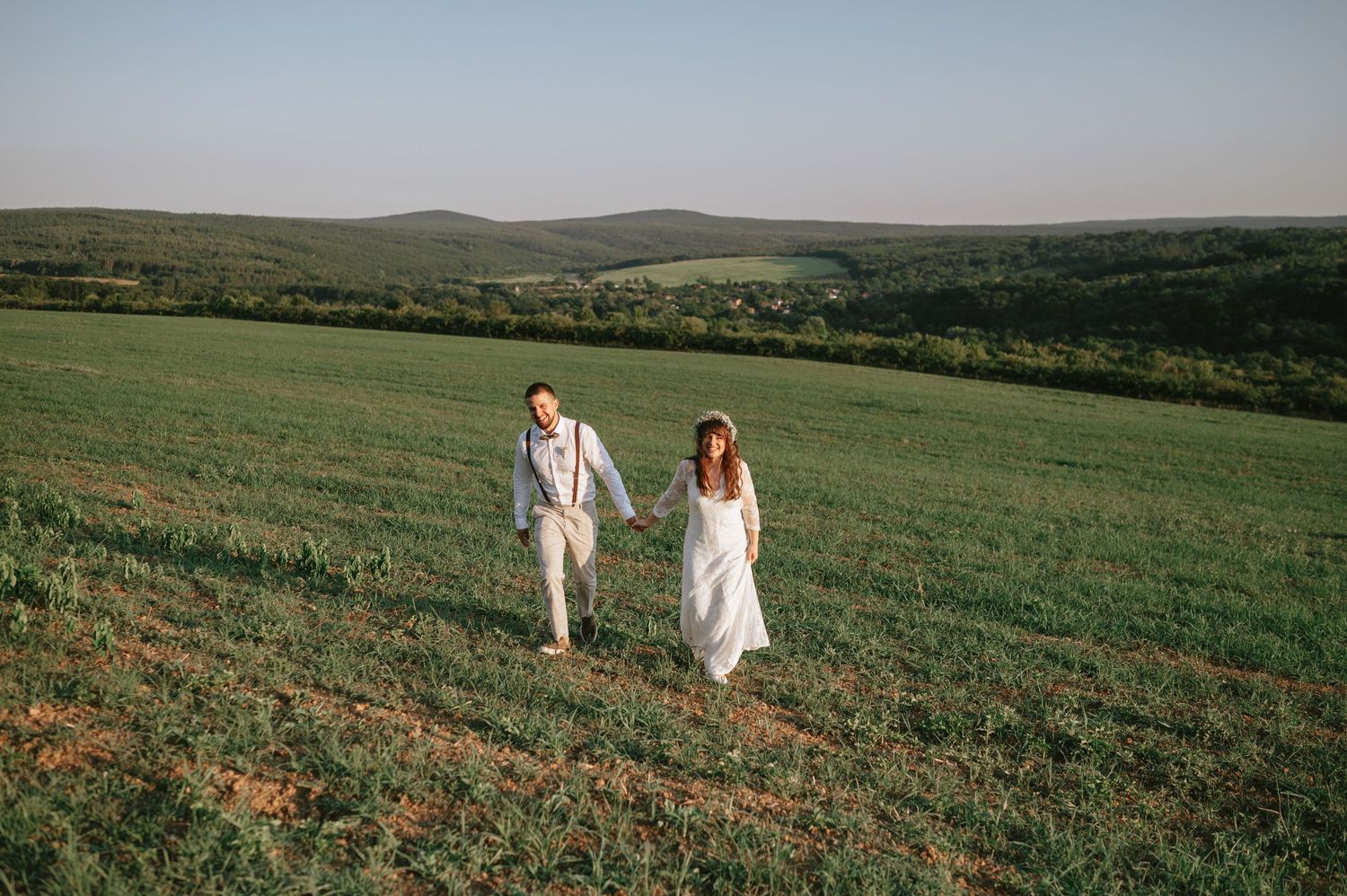Two people in wedding attire stroll through a scenic hillside meadow.