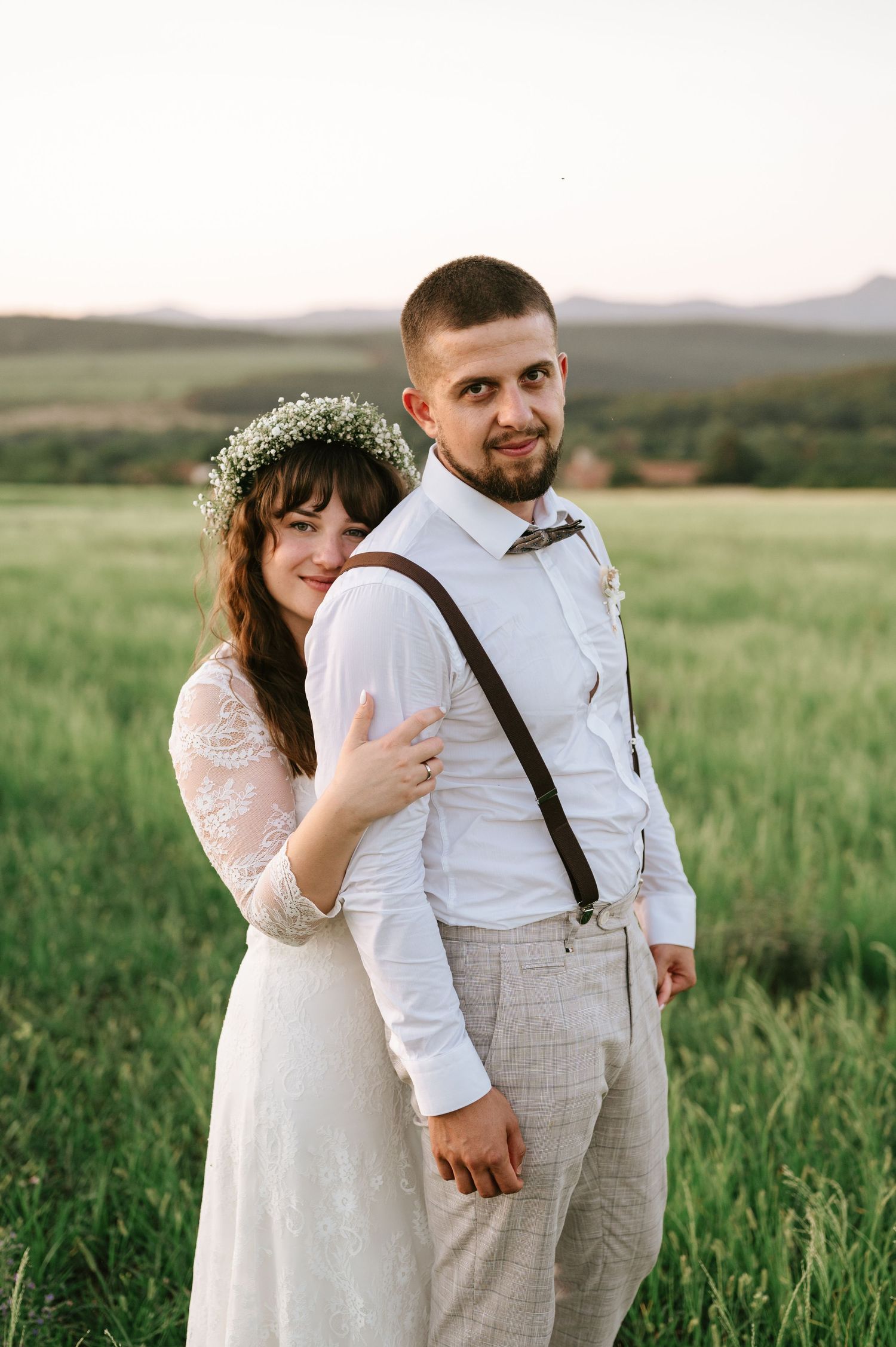 A couple in rustic wedding attire poses together in a green mountain meadow at sunset.