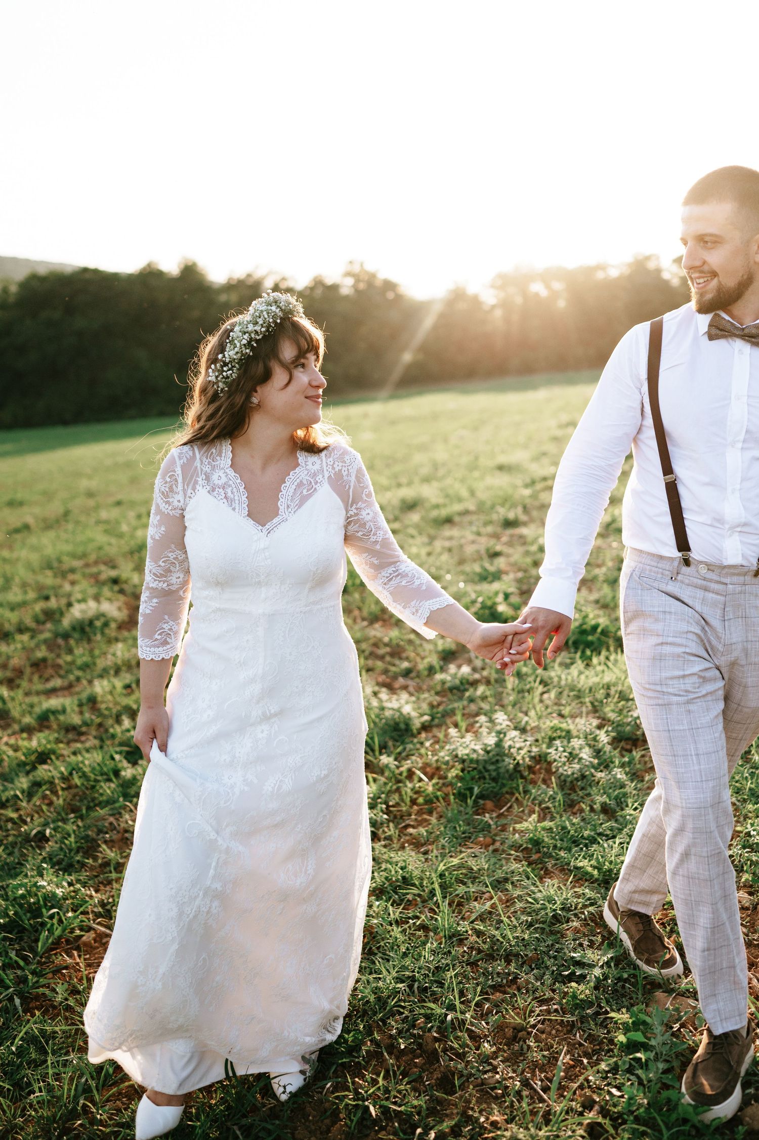 Wedding couple walking hand in hand through sunlit field in rustic outdoor setting.